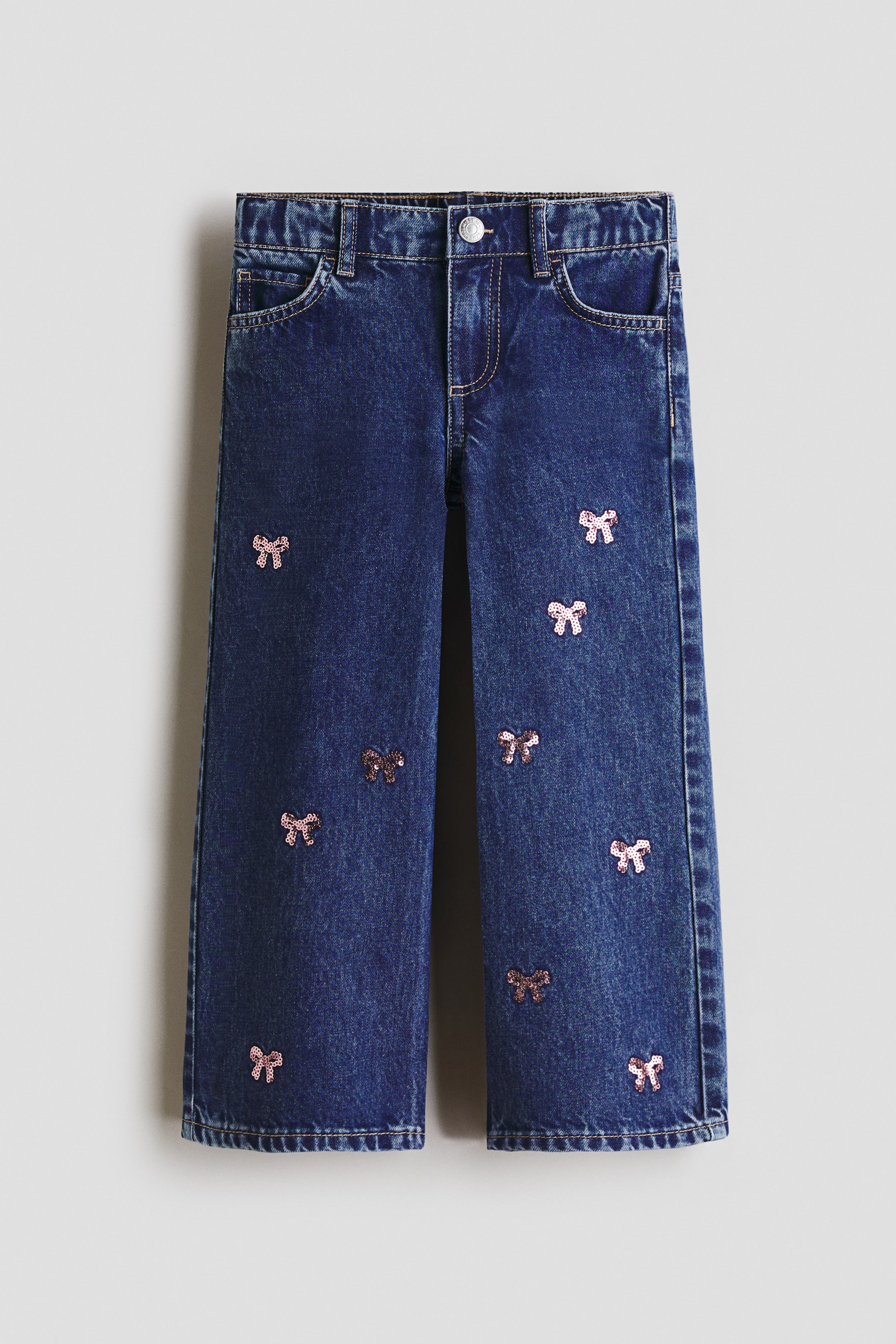 View larger image: Dark blue denim jeans featuring a button and zip closure, belt loops, front pockets with a coin pocket, and scattered pink sequin bow appliqués across both legs.