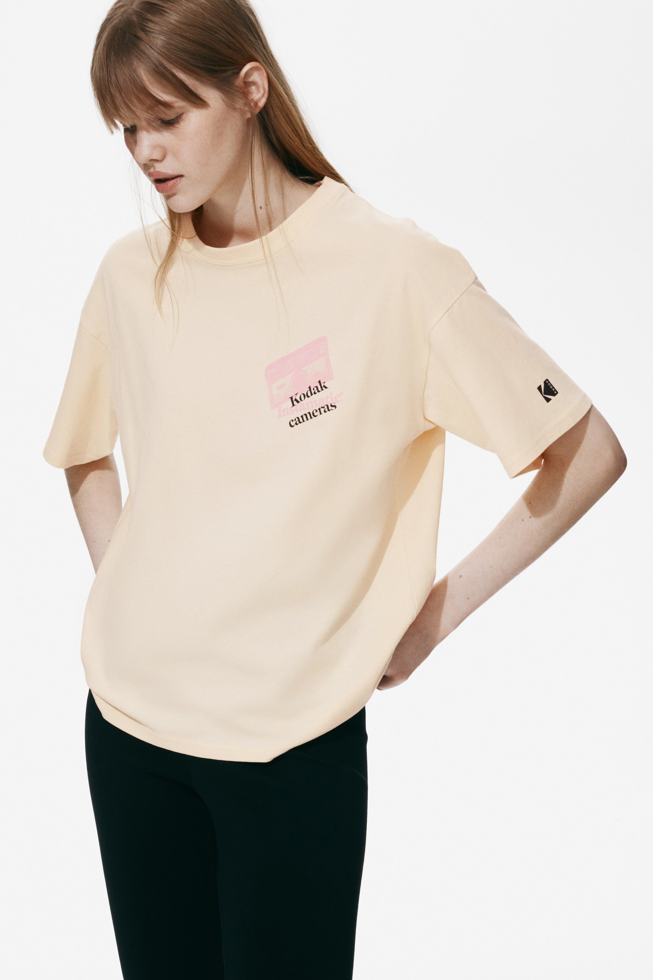 拡大画像を表示: 淡い黄色の半袖Tシャツは、左胸に「Kodak Instamatic cameras」と書かれたピンクのカメラのグラフィックが特徴で、左袖には小さな黒いKodakロゴが付いています。リラックスしたフィット感を持ち、やや斜めから見た前面ビューで表現されています。