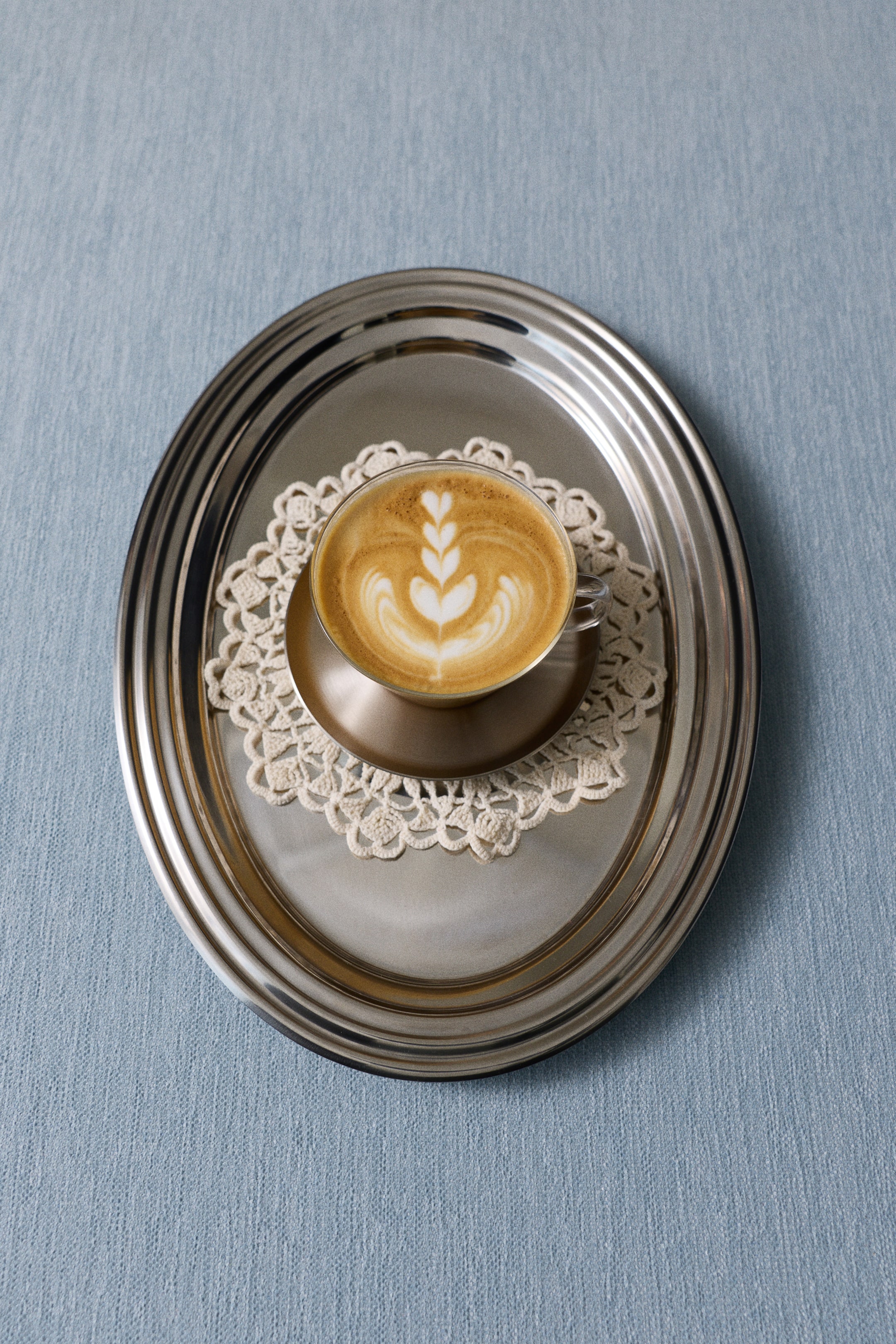 Visa större bild: En klar kaffekopp i borosilikatglas som innehåller en latte med hjärtformad skumdekoration vilar på ett runt underlägg i rostfritt stål med borstad finish. Koppen och underlägget är placerade på en vit virkad spetsdoily inuti en större oval bricka i rostfritt stål, sedd uppifrån.