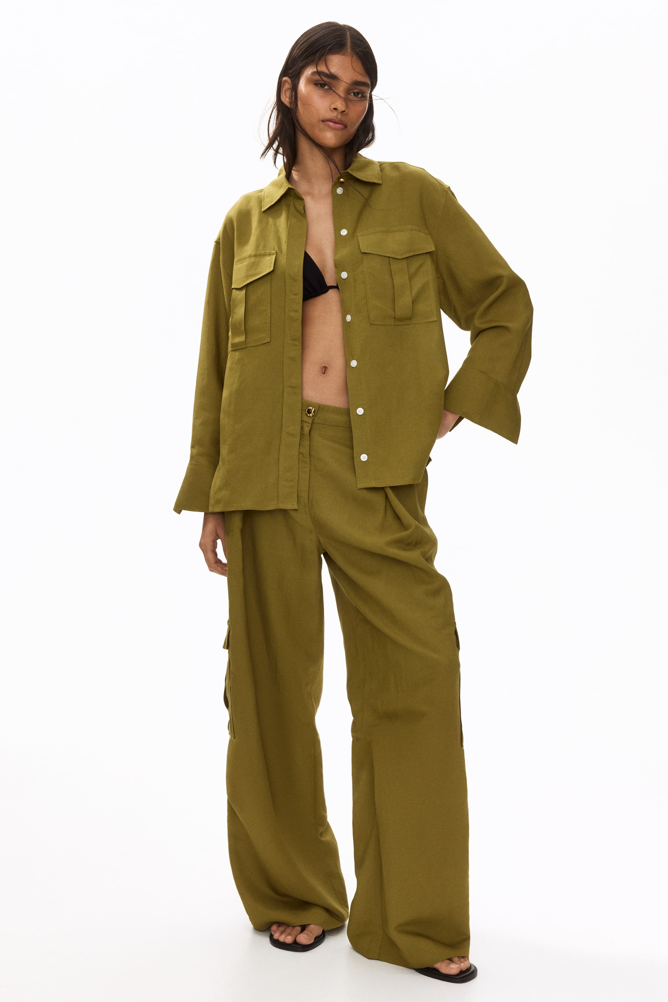View larger image: The olive green matching set is facing front, showcasing an oversized collared shirt with two chest flap pockets and wide cuffs, worn open over a black bra, and wide-leg pleated trousers with a visible cargo pocket and black sandals peeking from underneath.