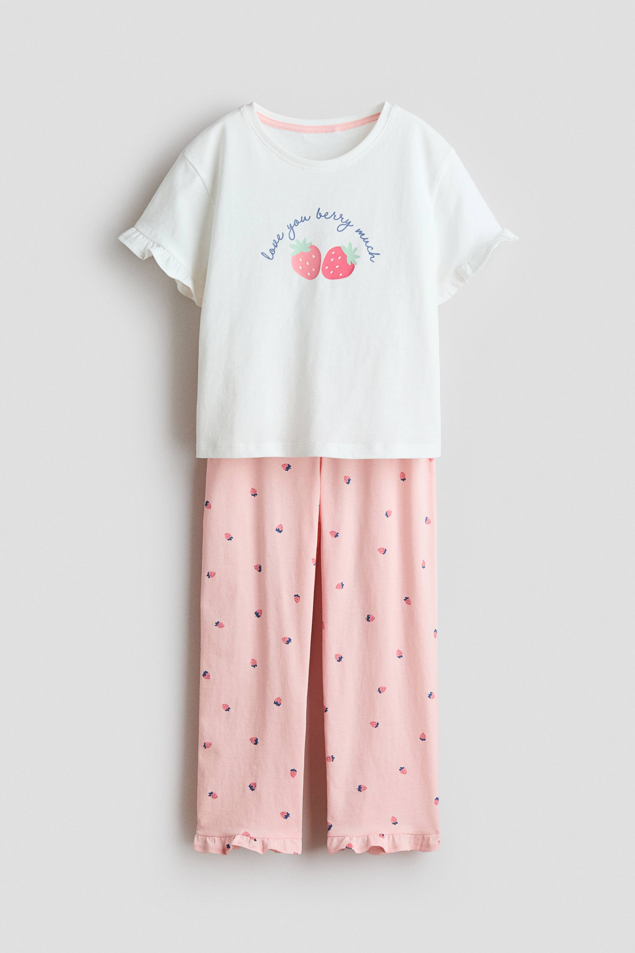 Se større billede: Et todelt pyjamassæt præsenterer sig fra forsiden, bestående af en hvid kortærmet top og lyserøde lange bukser. Toppen har teksten 'love you berry much' med to jordbærgrafikker og flæseærmer, mens bukserne viser et spredt jordbærprint og flæsemanchetter ved anklerne.