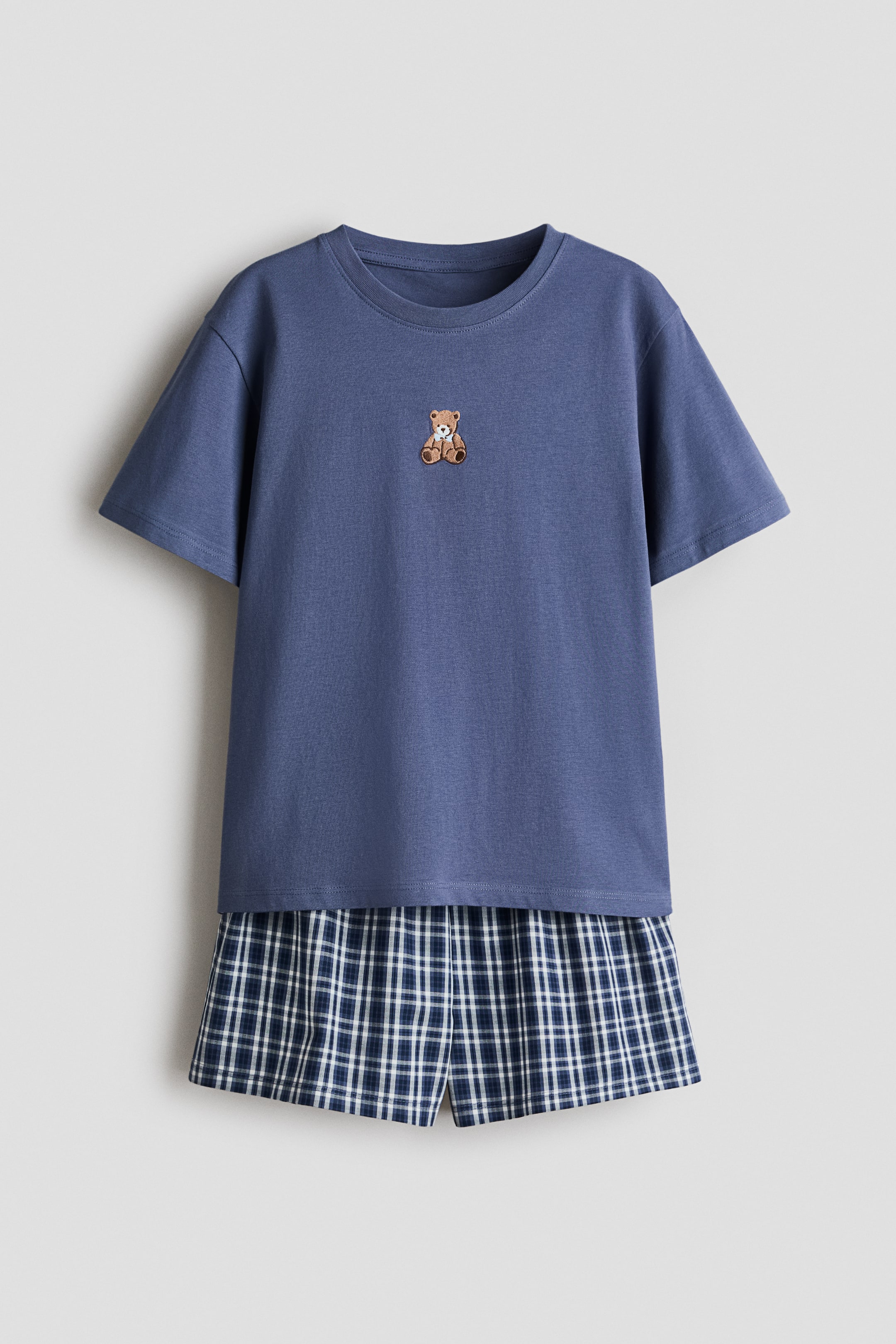 Agrandir l'image: Un ensemble pyjama présente un t-shirt bleu, avec un col rond à bord-côte, un petit motif d'ourson sur le devant et des épaules tombantes. Le t-shirt est superposé à un short à carreaux bleu, blanc et vert avec une taille élastiquée.