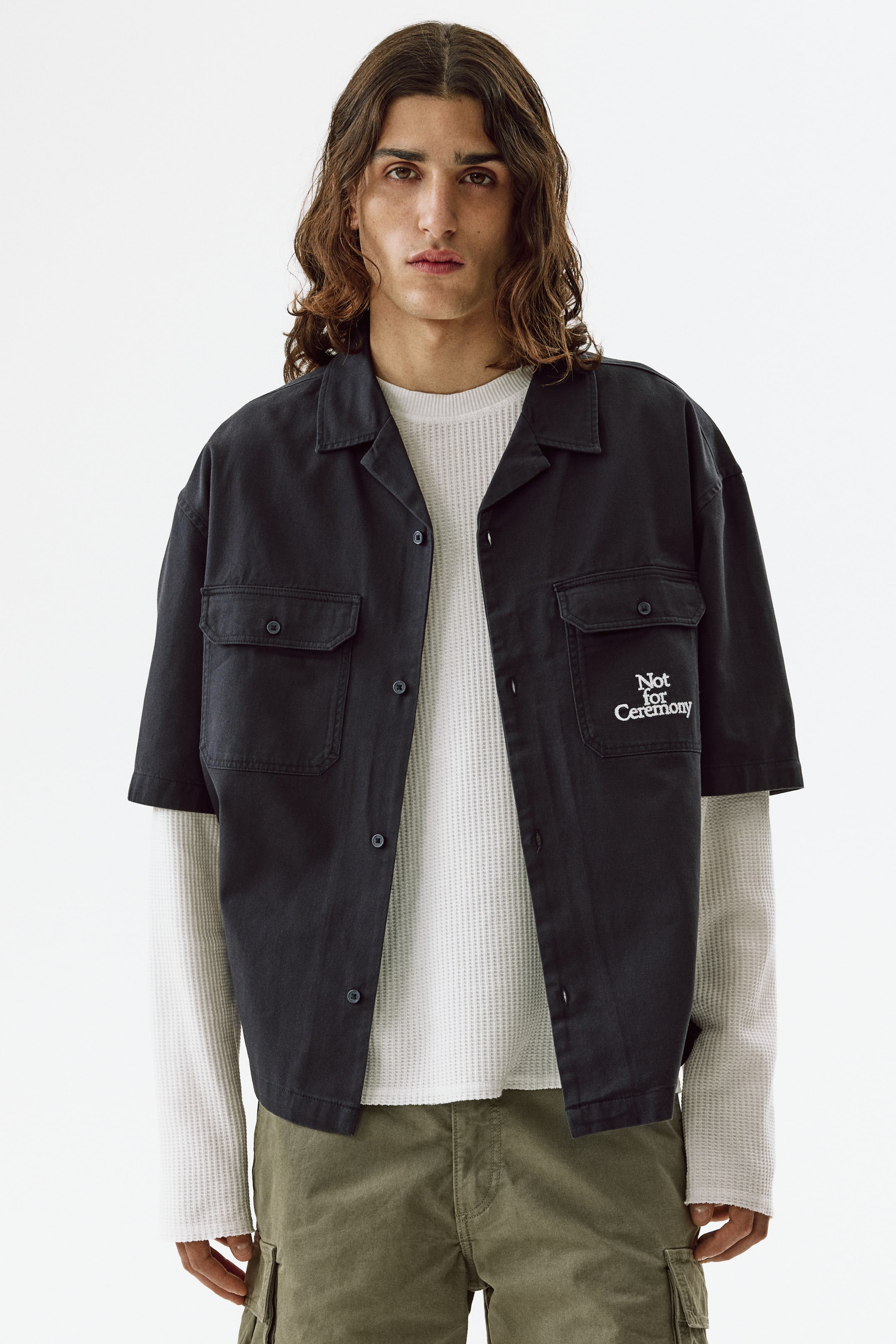 View larger image: A dark short-sleeved shirt features a revere collar, a button-down front with dark buttons, and two chest pockets with buttoned flaps. The right chest pocket displays white embroidered text reading "Not for Ceremony," and the shirt is worn open over a white waffle-knit long-sleeve shirt.