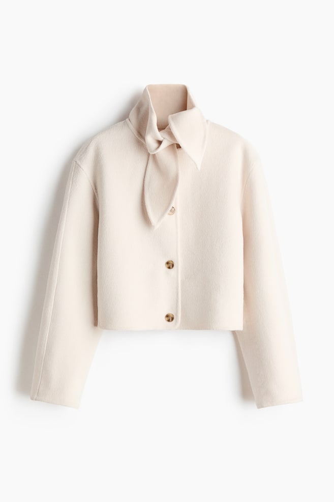 Une veste courte de couleur crème en mélange de laine feutrée se présente de face. Elle est dotée d'un col écharpe noué au cou, de trois boutons marron sur le devant et d'épaules légèrement tombantes. - 1