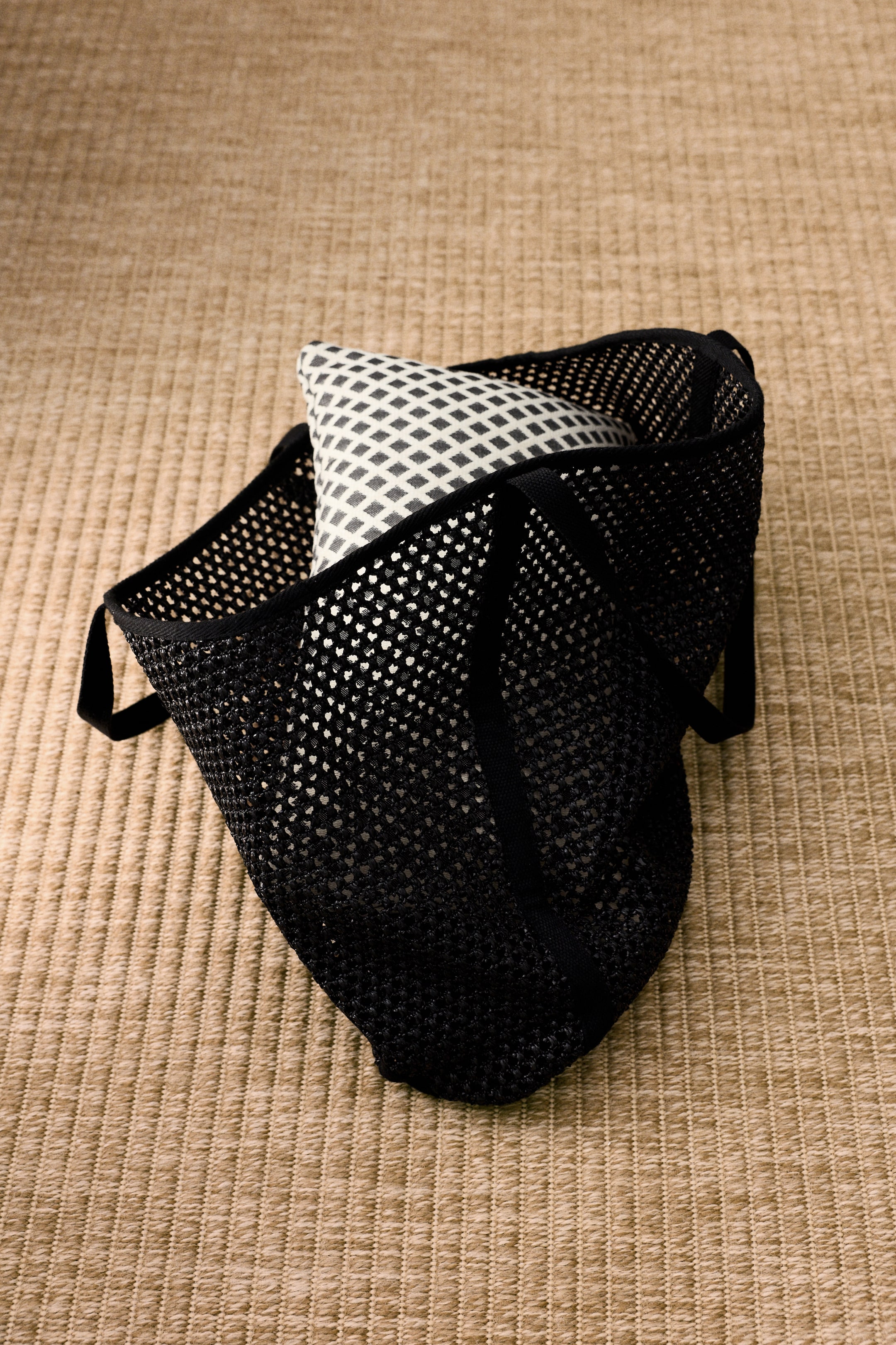 View larger image: A black mesh beach bag, crafted from plastic straw, is showcased with its open top, two handles, and sturdy, structured base from an angled perspective. Inside, a white pillow with a black geometric pattern rests, filling the bag's volume.