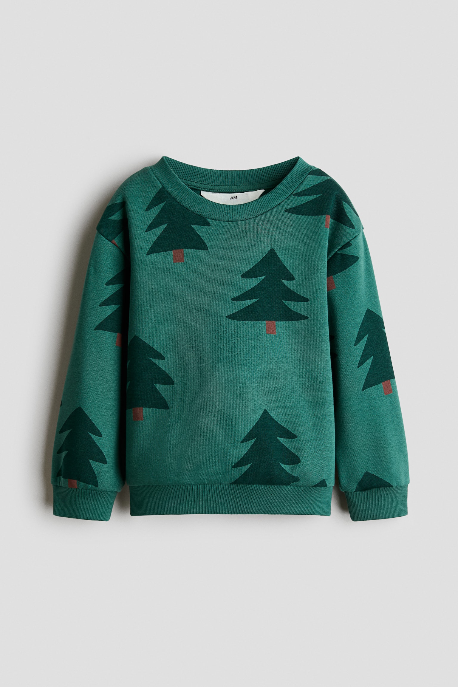 A green sweatshirt is shown from the front, featuring a printed design of dark green pine trees with brown trunks. The sweatshirt includes a round ribbed neckline, dropped shoulders, long sleeves with ribbed cuffs, and a ribbed hem.