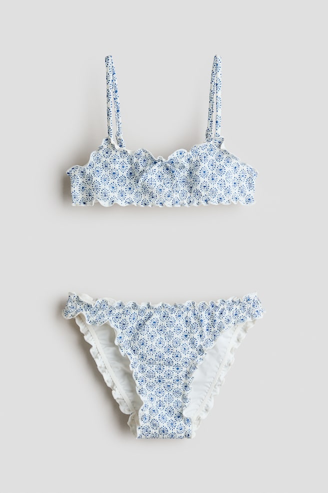 Een vooraanzicht van een bikiniset toont een blauw-wit patroon op een top met gesmokt materiaal, smalle verstelbare schouderbandjes en een ingenaaid strikje aan de voorkant, gecombineerd met bijpassende gesmokte broekjes; beide stukken hebben geschulpte overlockranden. - 1