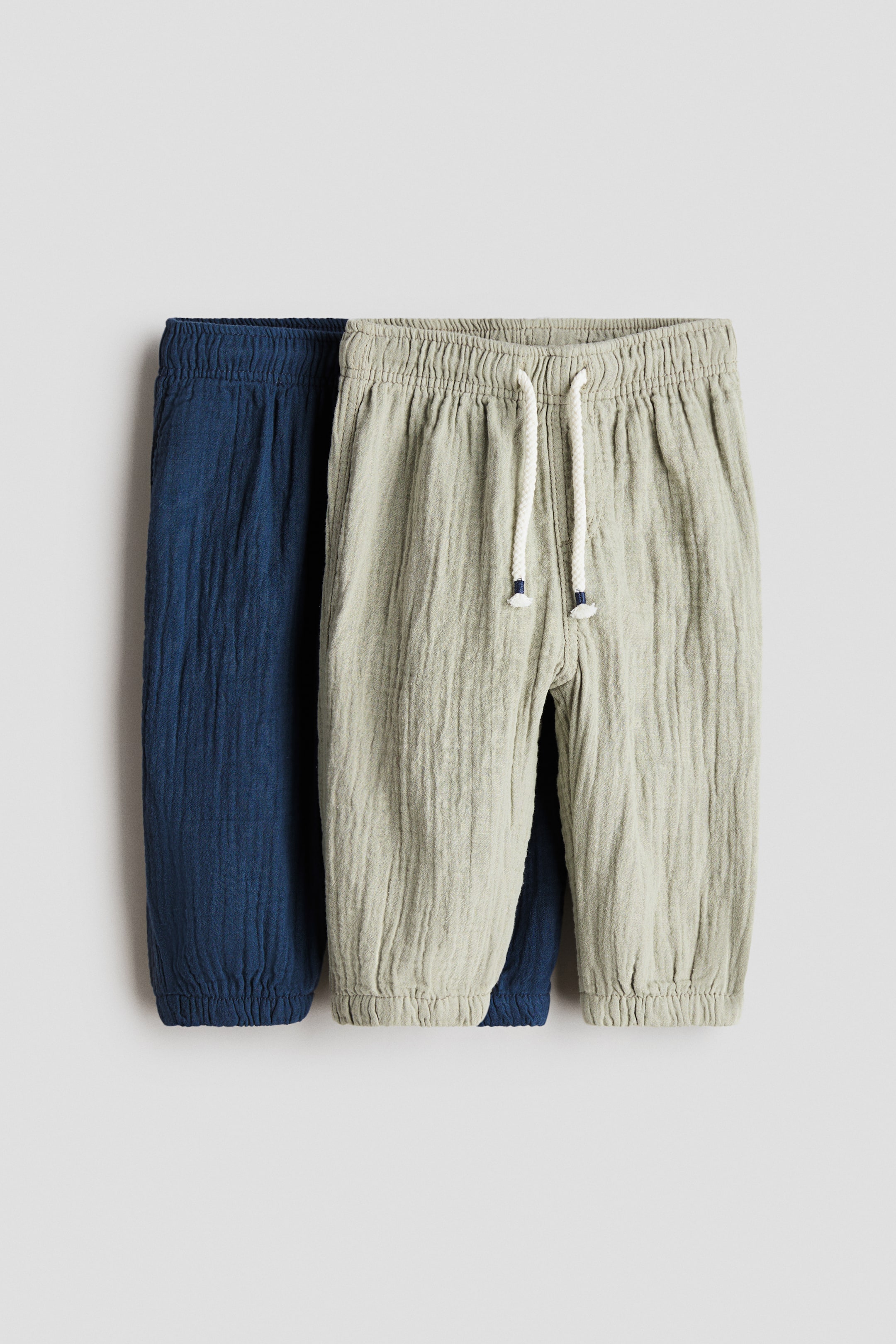 View larger image: Two pairs of trousers are front-facing, both with a crinkled texture and elasticated ankle cuffs. The light-green pair features an elasticated waistband with a white drawstring tipped in blue, while the dark-blue pair features an elasticated waistband.