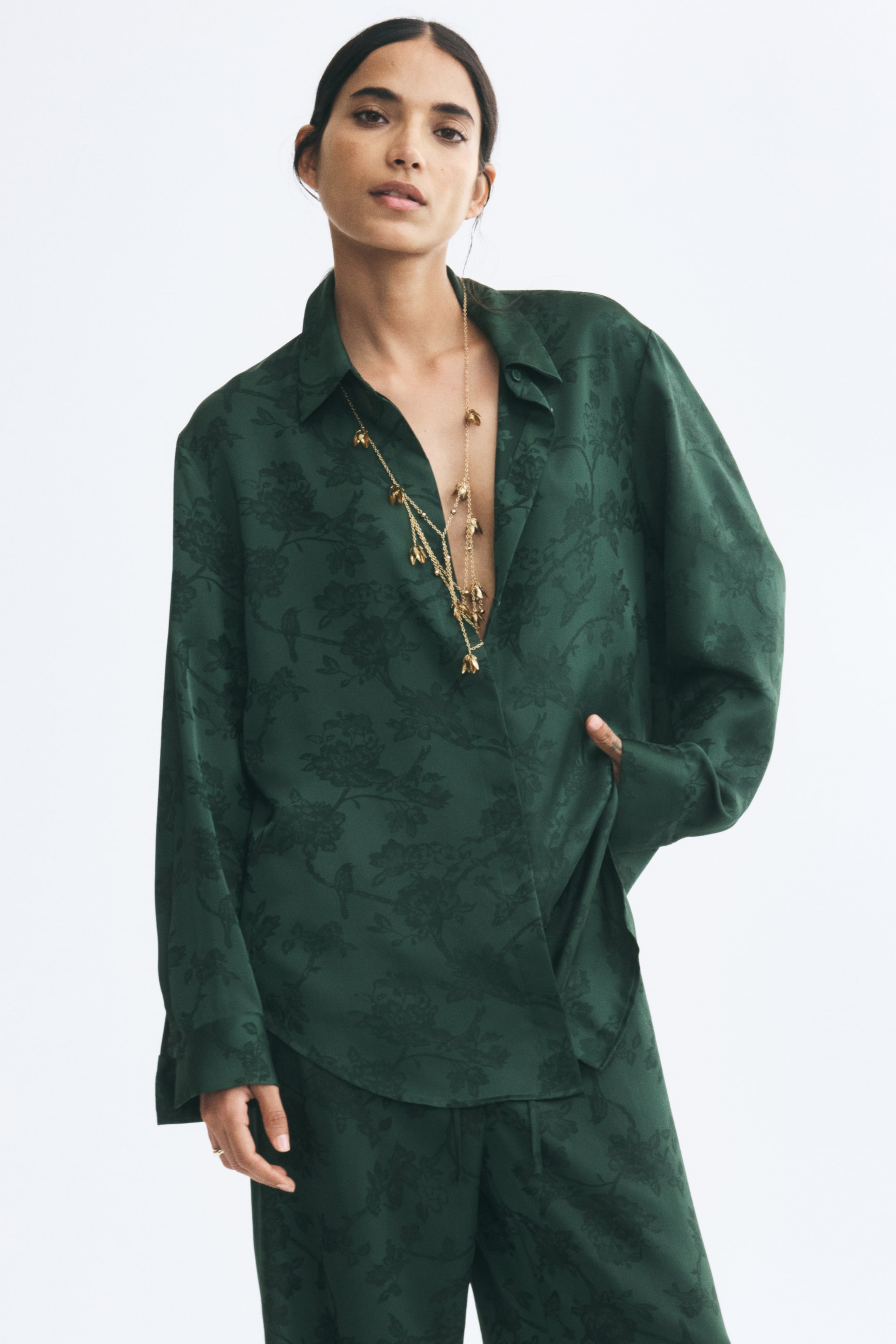 View larger image: A long shirt in a dark green floral jacquard weave is being worn by the model, featuring a collar, concealed front buttons, and long sleeves with button cuffs. The shirt is styled open at the collar with layered gold necklaces and paired with matching jacquard trousers.