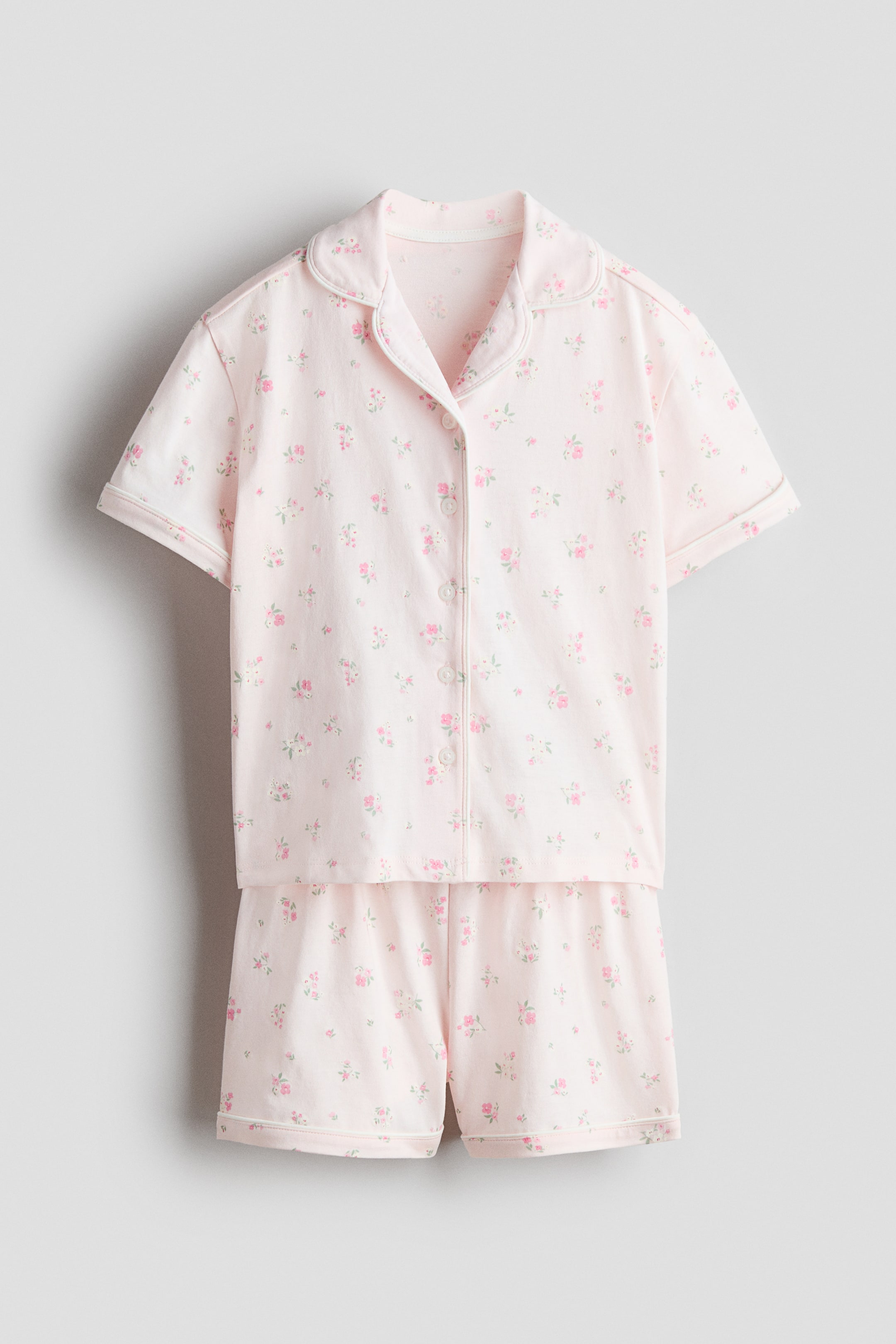 Büyük resmi görüntüle: Önden bir görünüm, açık pembe kısa kollu bir gömlek ve uyumlu şorttan oluşan desenli pijamaları gösteriyor; her iki parça da küçük çiçek desenine sahip. Gömlek resort yakalı, önden düğmeli ve düşük omuzlu olup yaka, düğme bandı ve kol ağızlarında beyaz biyeye sahiptir. Şortlar ise lastikli bele ve paça uçlarında uyumlu beyaz biyeye sahiptir.