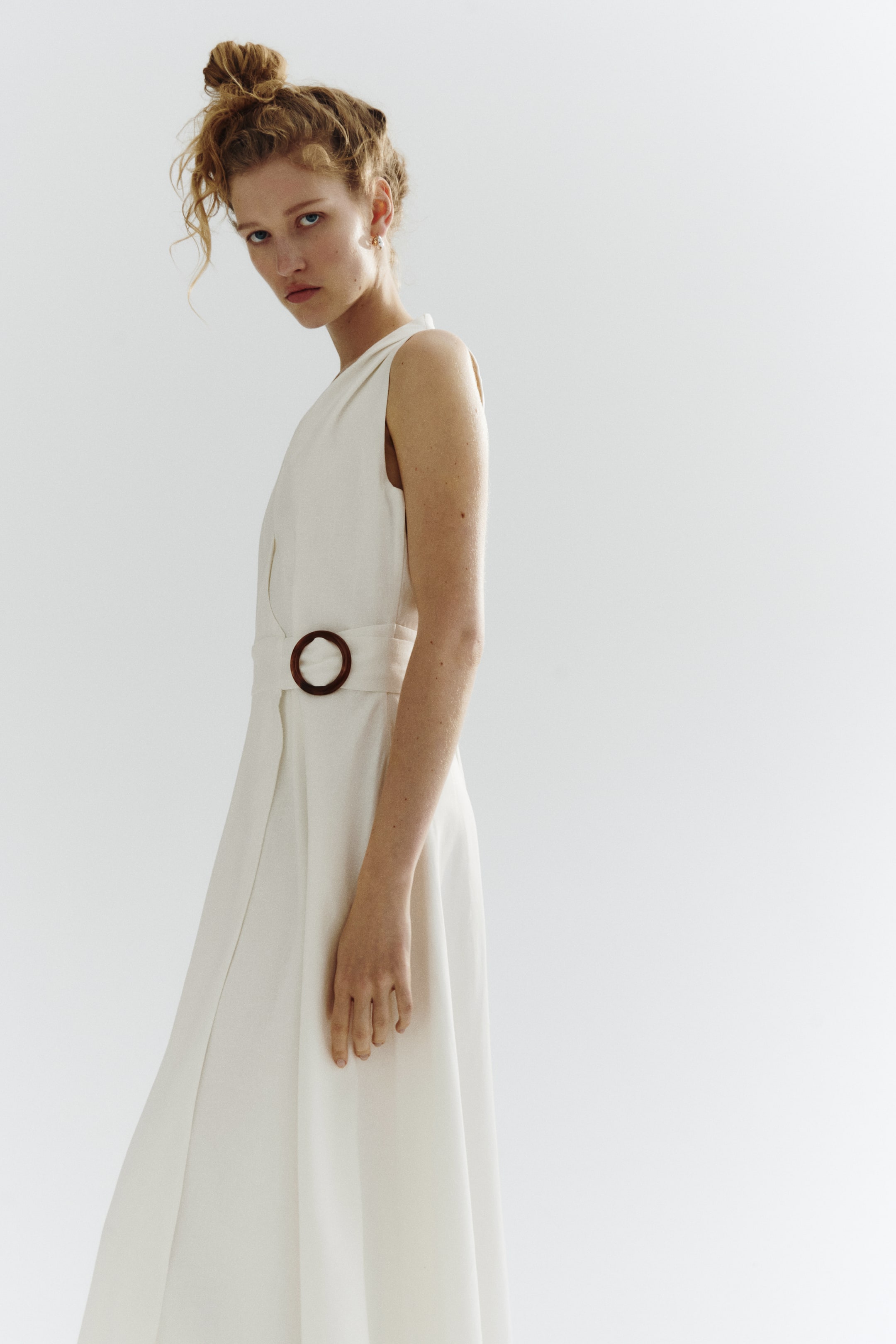 View larger image: An off-white sleeveless wrap dress features a V-neckline. A wide self-fabric belt cinches the waist with a round, dark brown buckle, and the dress flows into a long skirt.