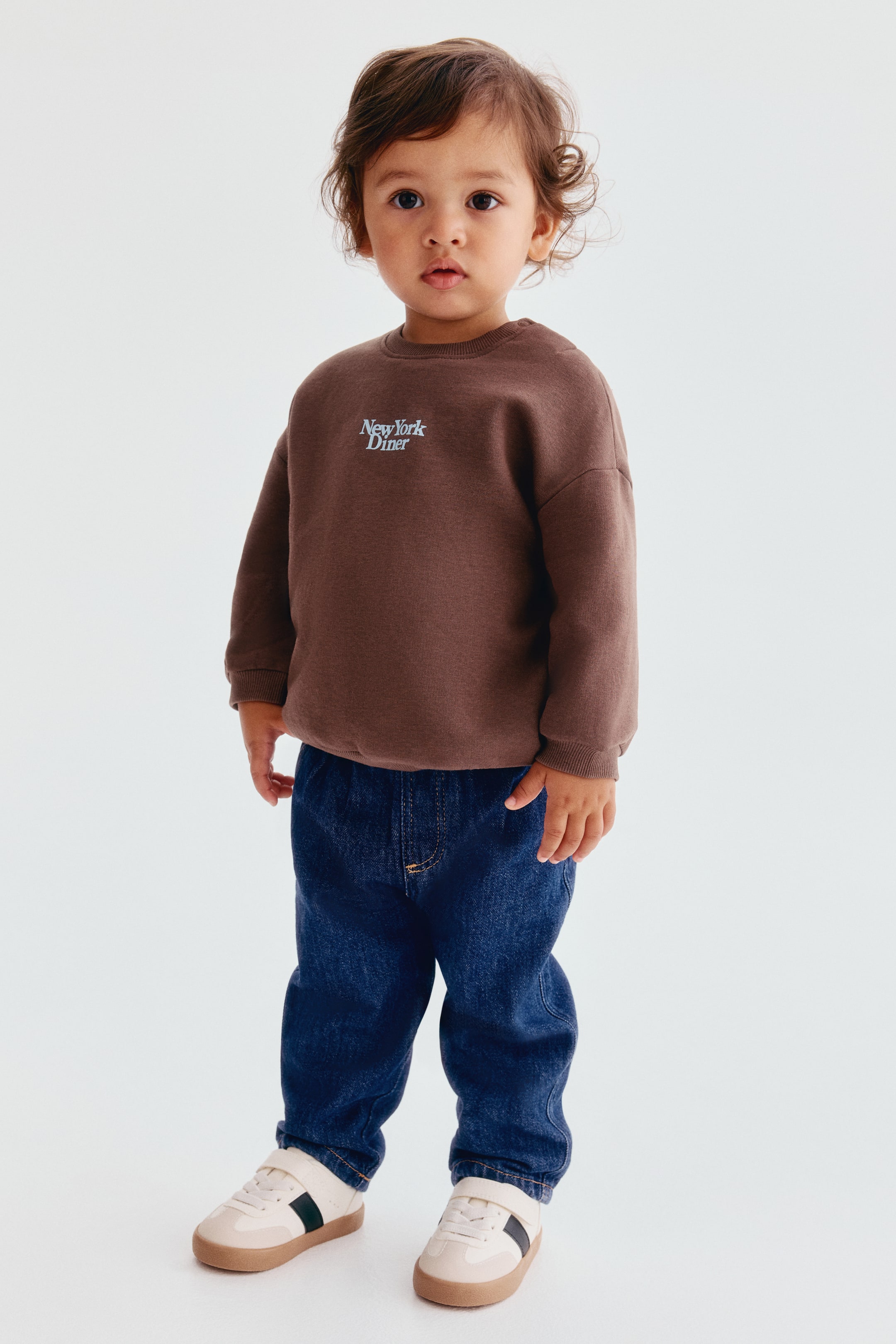 View larger image: This sweater and joggers set, shown from the front, features a brown crew-neck sweatshirt with dropped shoulders, ribbed cuffs, and "New York Diner" printed in white text on the chest. The sweatshirt is paired with dark blue, denim-look joggers featuring a relaxed fit, an elastic waistband, and contrasting topstitching.