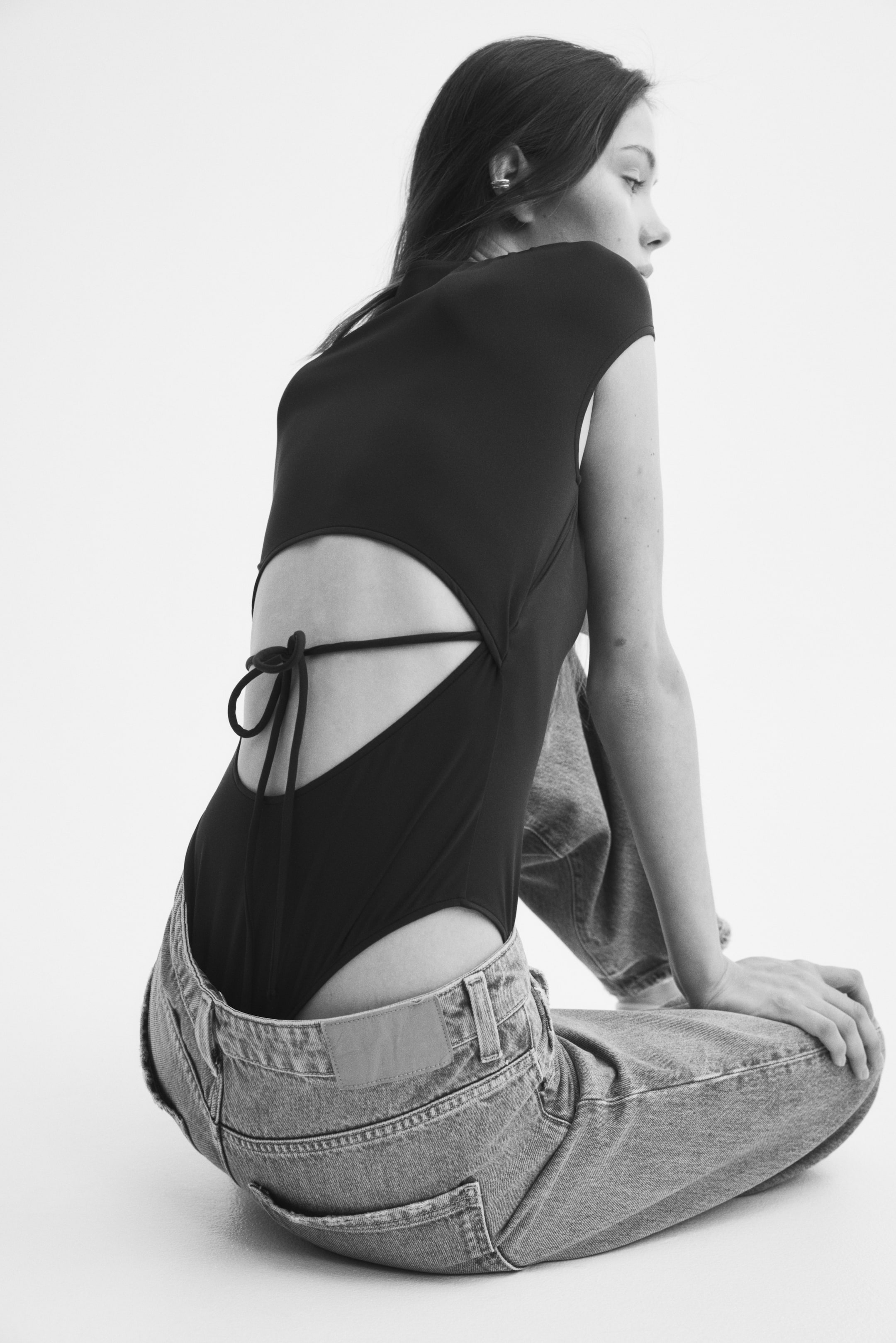 View larger image: A black stretch-jersey bodysuit with cap sleeves and a large cut-out section at the back with narrow ties is being worn by the model. The model is seated facing away from the camera, showcasing the bodysuit's back details while wearing it layered under light-wash denim jeans.