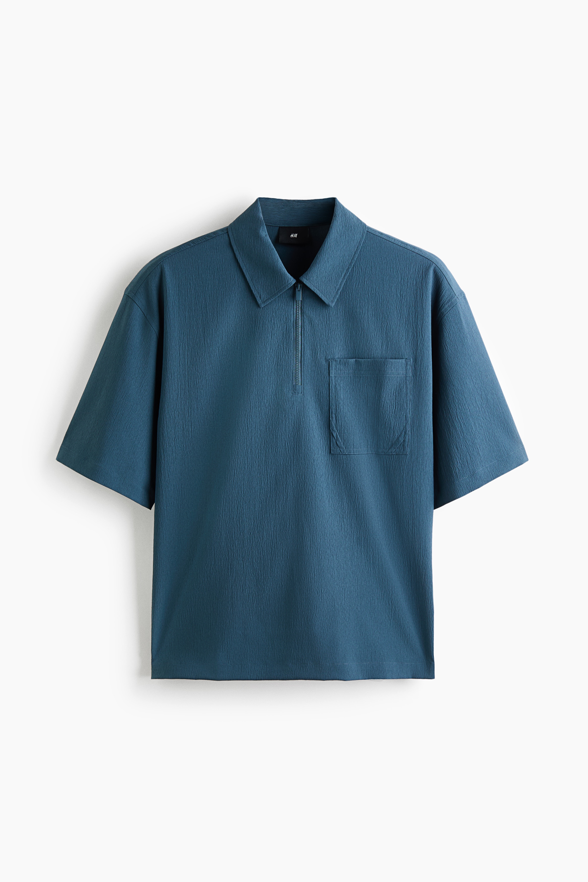 The model is wearing a blue polo shirt in a crinkled weave, featuring a relaxed fit, a collar with a partially open half-zip, short sleeves, and a single chest pocket. The shirt is layered over a white T-shirt and paired with grey trousers.