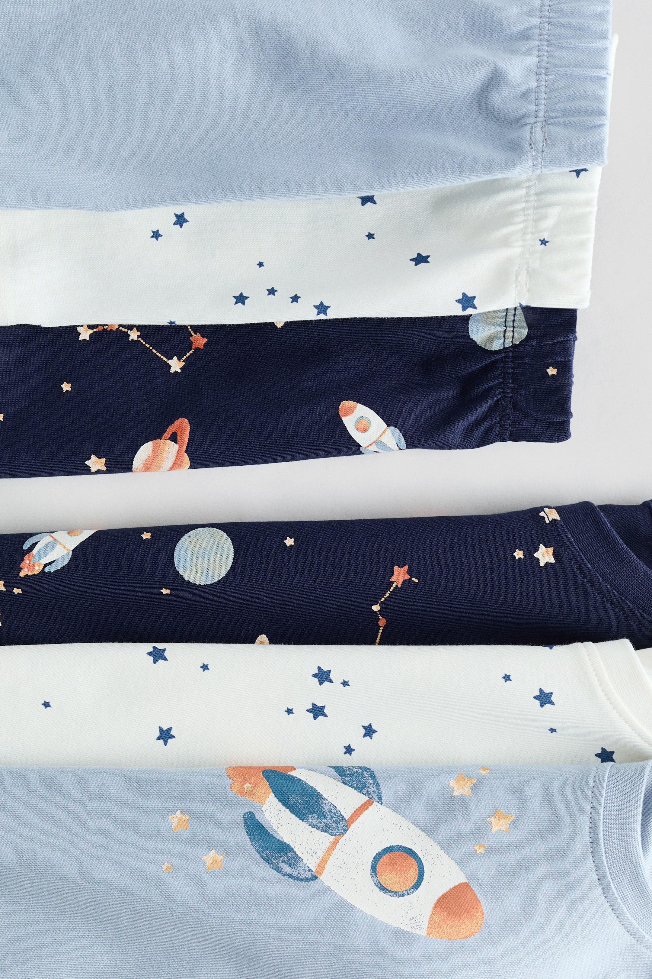 Größeres Bild anzeigen: Ein 3er-Pack kurze Baumwollpyjamas zeigt drei verschiedene Designs, flach ausgelegt. Die Sets bestehen aus einem hellblauen Stoff mit Raketen- und Sternenmuster, einem weißen Stoff mit verstreuten blauen Sternen und einem dunkelblauen Stoff mit Illustrationen von Raketen, Planeten und Sternbildern. Elastische Taillenbänder und gerippte Halsausschnitte sind an den verschiedenen Teilen sichtbar.