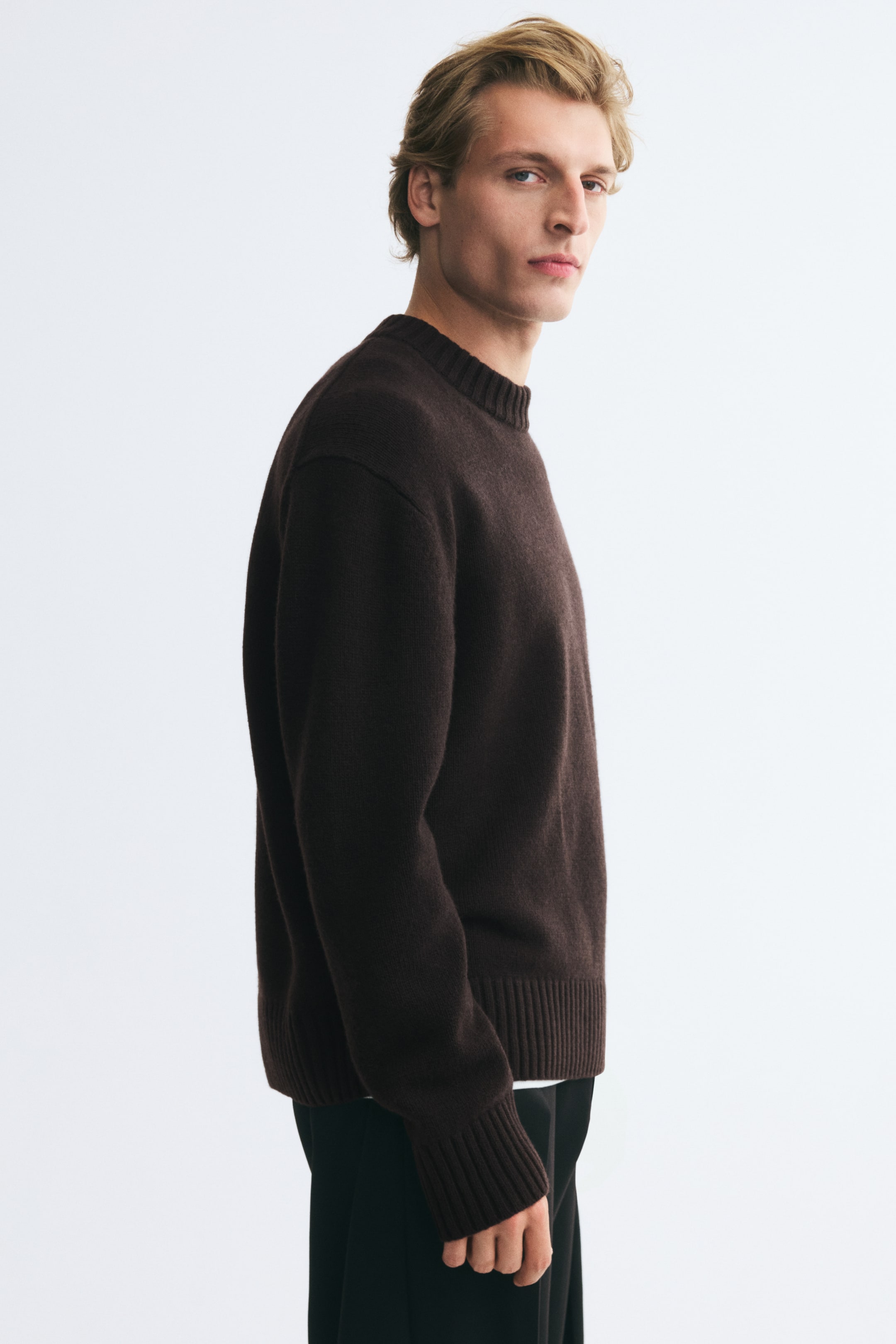 View larger image: The model is wearing a dark brown knit sweater with a ribbed crew neck, long sleeves, and ribbed cuffs, paired with black trousers.