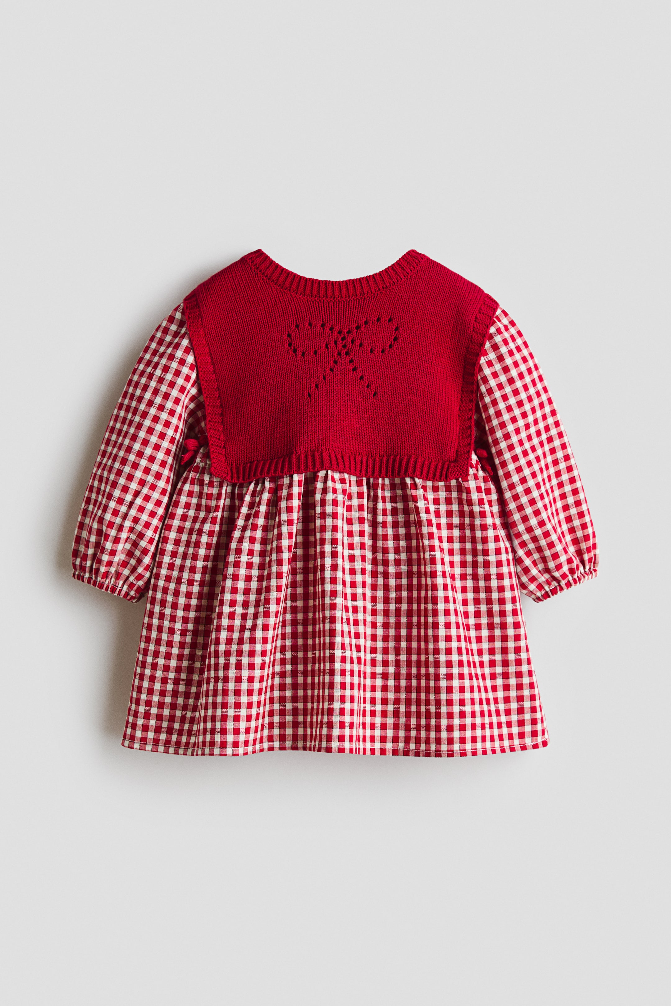 View larger image: A child’s dress is shown from the front, featuring a red knitted upper section with an openwork bow design, combined with a red-and-white gingham check lower section and puffy long sleeves gathered at the cuffs.