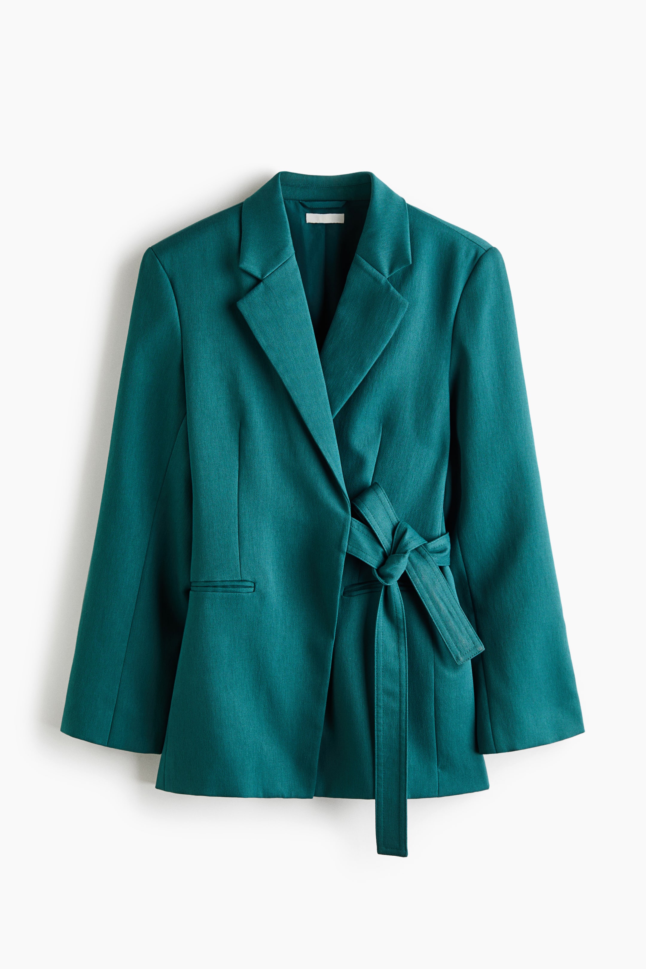 Büyük resmi görüntüle: Önü dönük olan petrol yeşili kruvaze blazer, yırtmaçlı yakaları, yapılandırılmış omuzları ve uzun kollarıyla öne çıkıyor. Blazer, bel kısmında yandan bağlanan düğümlü kendi kumaşından bir kuşakla kapanıyor ve ince welt cepleri içeriyor.