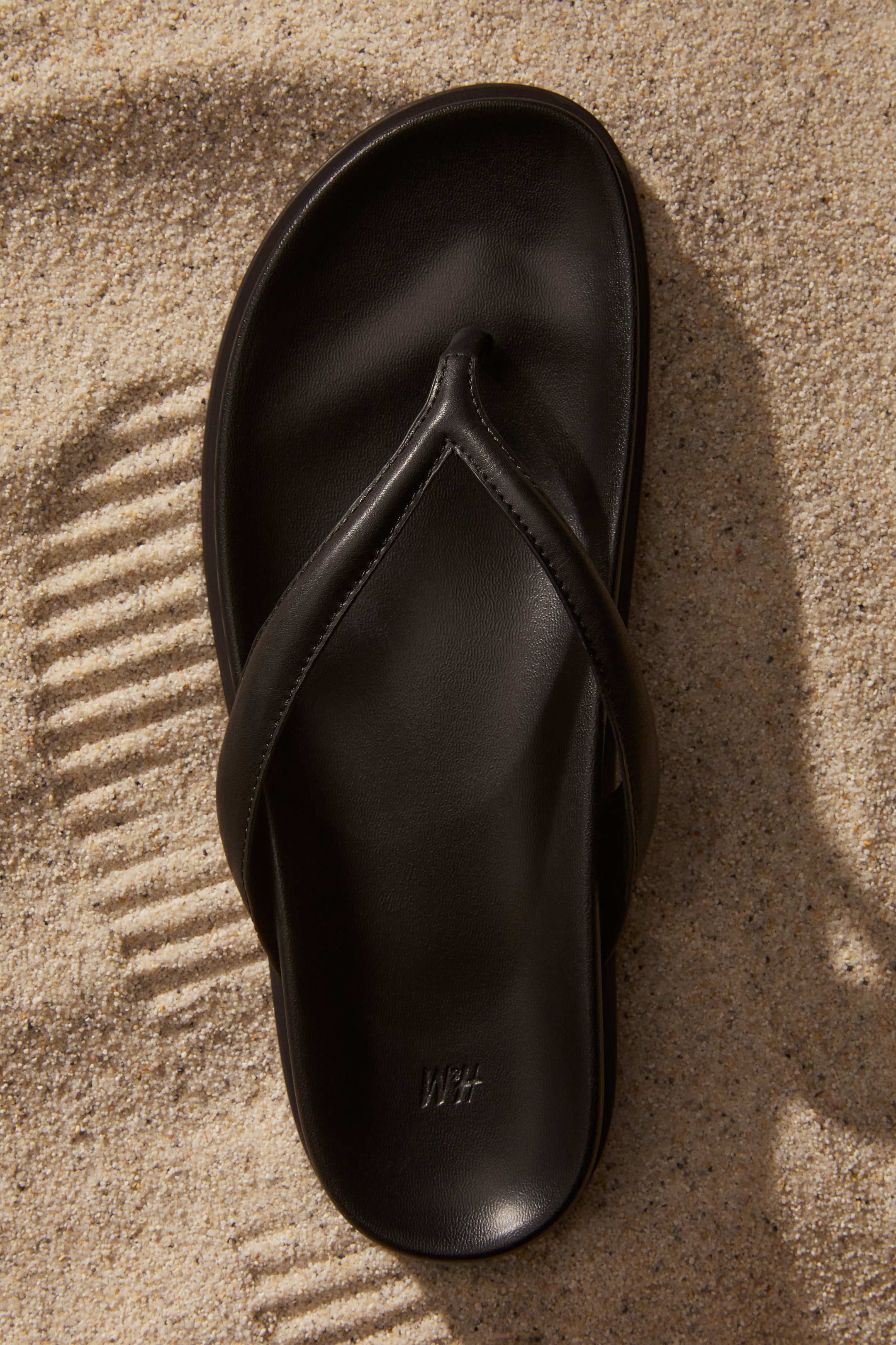 Vezi imaginea mai mare: Un șlapi negru este așezat plat, văzut dintr-o perspectivă de sus în jos, având o talpă netedă și mată cu o curea căptușită în stil thong, formând o formă de V. Logo-ul 'H&M' este debosat pe talpă, aproape de călcâi.