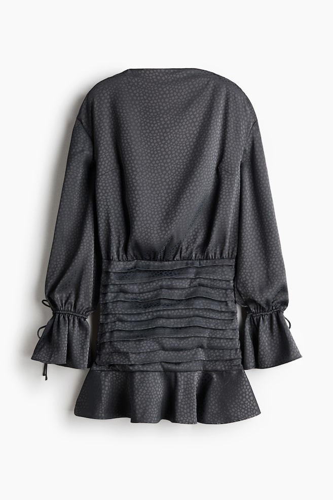 Un vestido corto se presenta de espaldas, con una tela oscura que presenta un patrón texturizado sutil en toda su superficie. Muestra un escote trasero redondeado y un cuerpo de corte holgado, con mangas largas y hombros caídos que se extienden hasta puños acampanados con cordones ajustables. El vestido exhibe una sección media fruncida horizontalmente, que fluye hacia una falda con un dobladillo con volantes. - 1