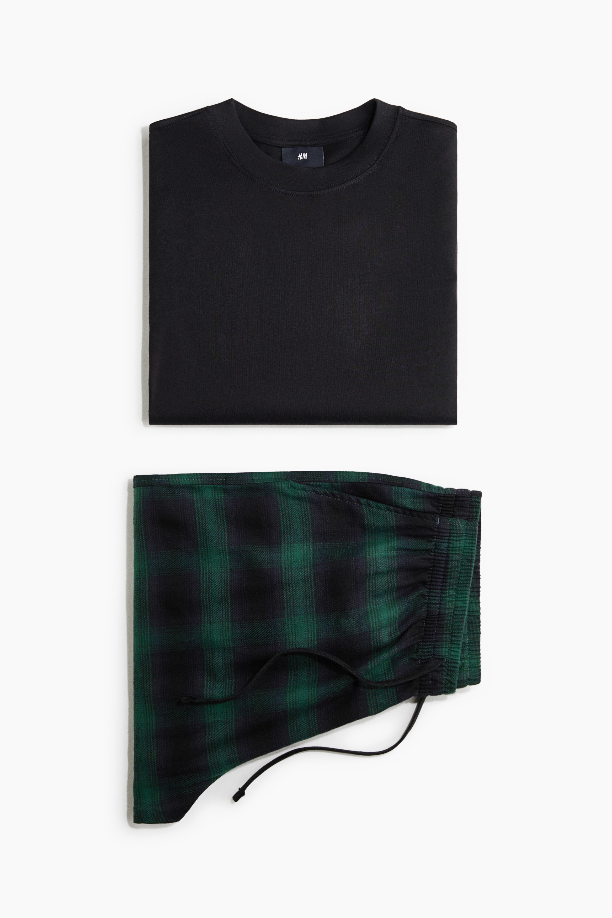 Een katoenen loungewear-set bestaande uit een zwarte jersey top met lange mouwen en verlaagde schouders, gecombineerd met een paar wijde flanellen broek in groen en zwart ruitdessin, wordt gedragen door het model. Het model staat met Ã©Ã©n hand in de broekzak, waardoor de relaxte pasvorm en de taille met trekkoord extra worden benadrukt.