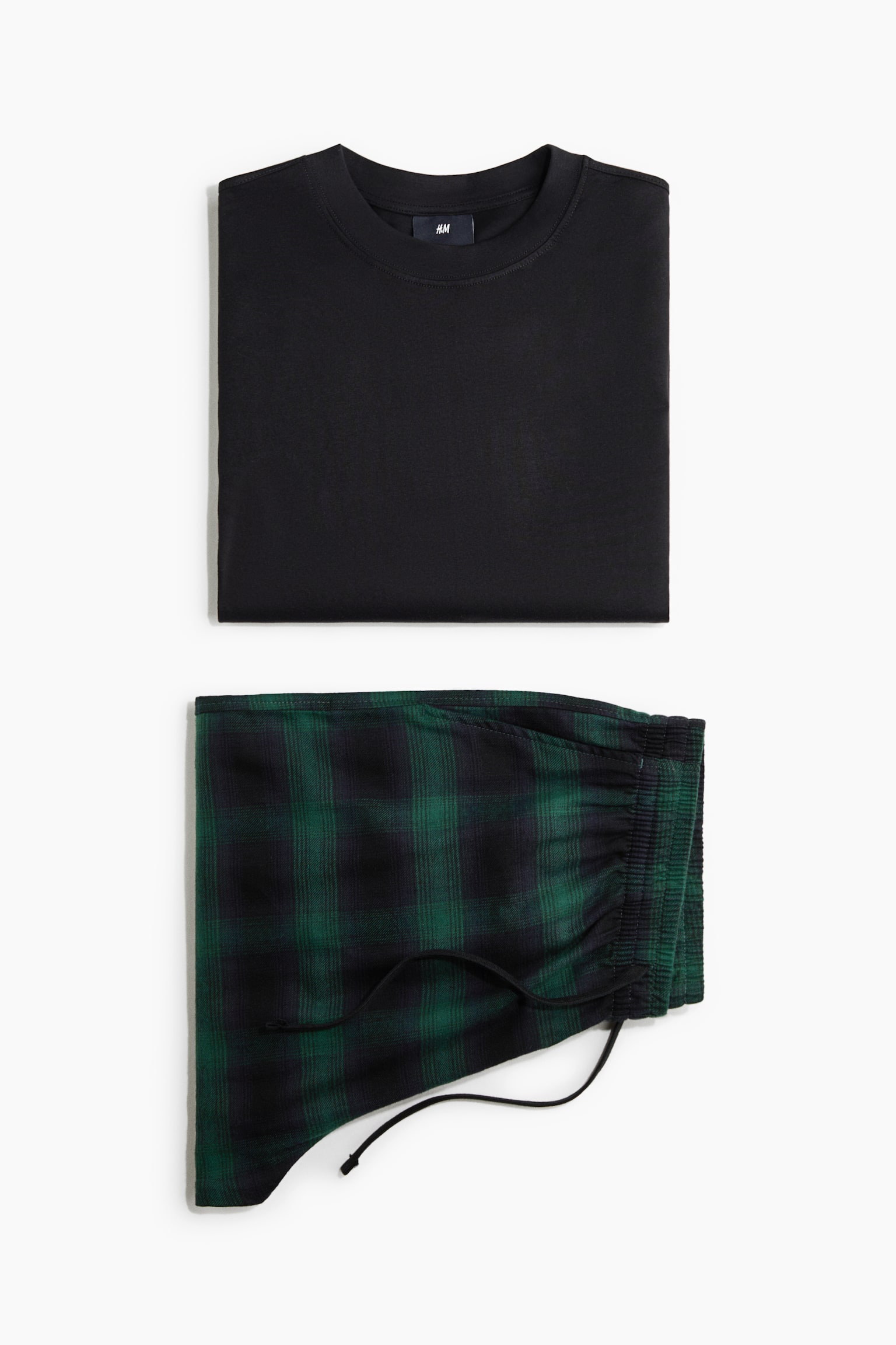 DwuczÄÅciowy zestaw do domu przedstawiono w ujÄciu flat lay, zÅoÅ¼ony z jednolitego czarnego T-shirtu z okrÄgÅym dekoltem uÅoÅ¼onego nad zÅoÅ¼onymi zielono-czarnymi flanelowymi spodniami piÅ¼amowymi w kratÄ. Spodnie pokazujÄ marszczony elastyczny pas z czarnym sznurkiem do ÅciÄgania. - 1