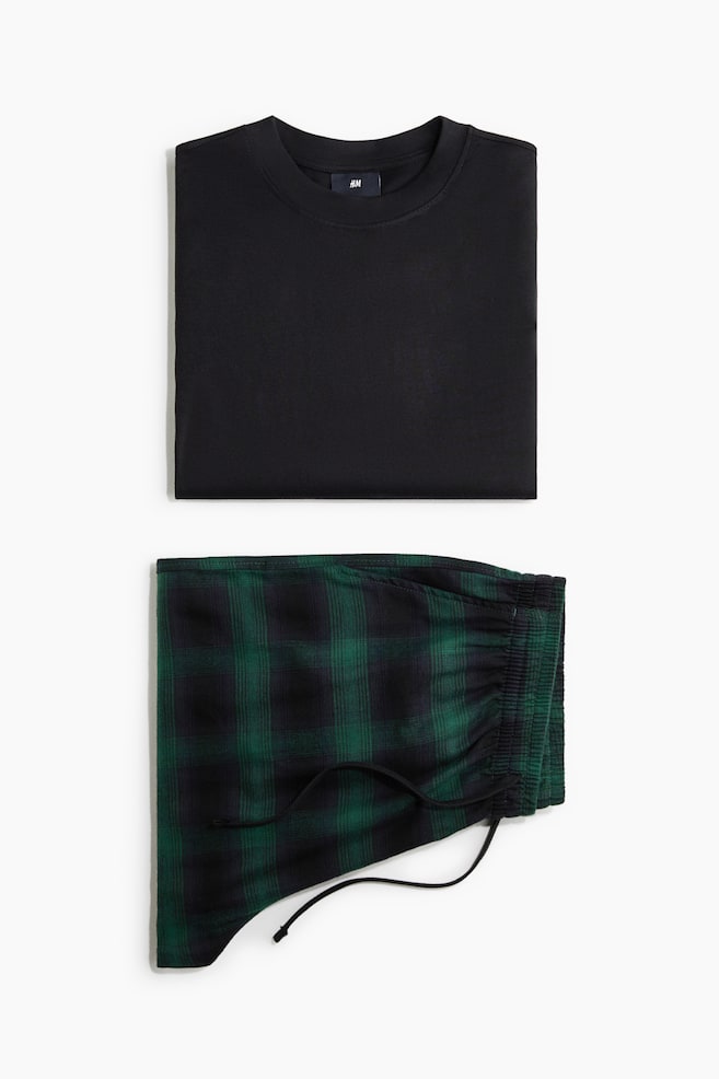 Et todelt lounge-sett presenteres i flat-lay-visning, med en foldet ensfarget svart crewneck T-skjorte plassert over foldede grÃ¸nne og svarte rutete pysjbukser. Buksene viser en rynket elastisk linning med svart snÃ¸ring. - 1