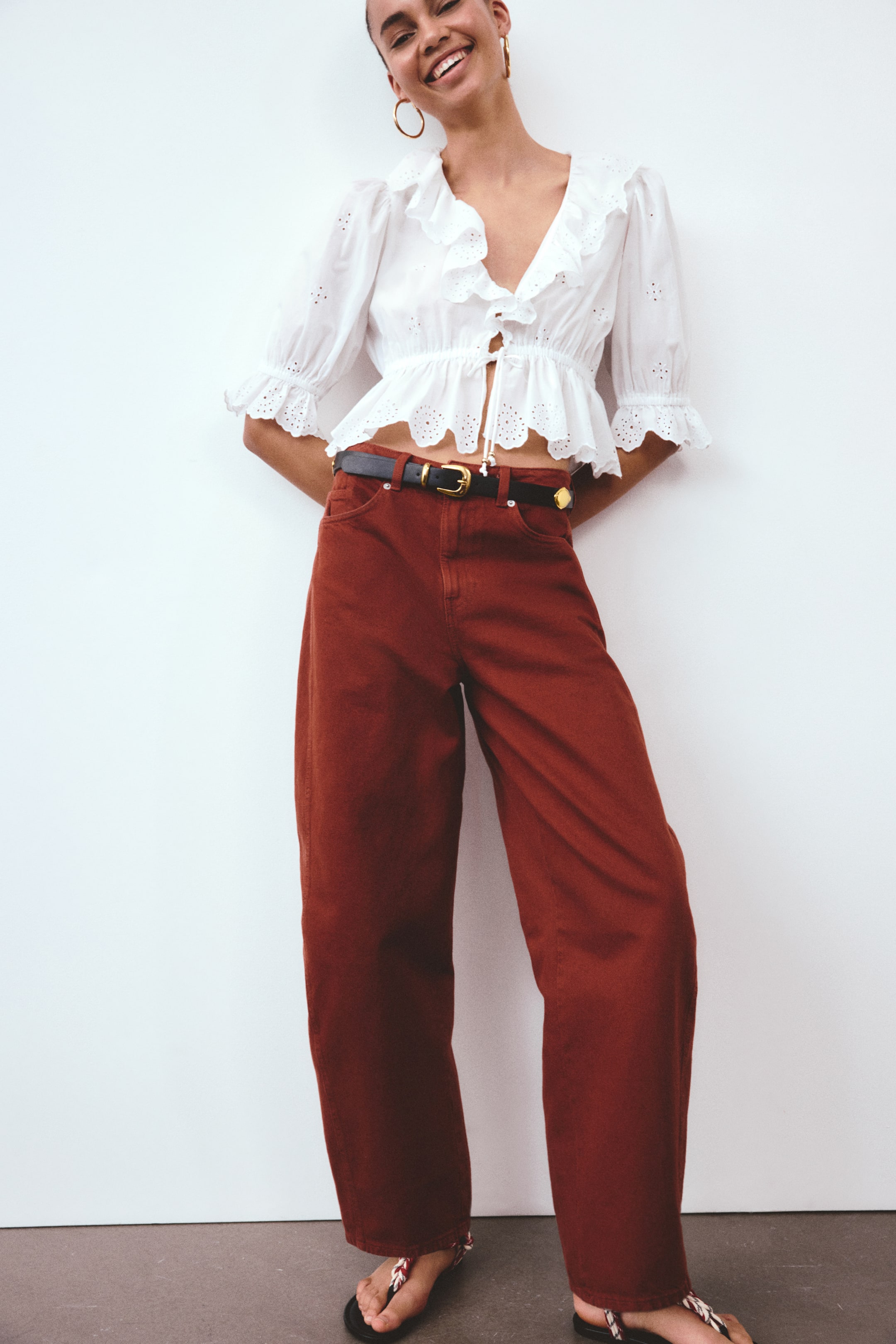 הצגת תמונה גדולה יותר: חולצת פפלום רקומה בלבן עם חורים, הכוללת מחשוף V עם עיטורים, שרוולים תפוחים, סגירה עם שרוך קשירה, ושוליים מסולסלים, נלבשת. היא משולבת עם מכנסיים בגזרה גבוהה ורחבים בצבע חום-אדמדם, שחגורה שחורה עם אבזם בגוון זהב מהדקת אותם, וסנדלים שטוחים עם רצועות קלועות משלימים את המראה.