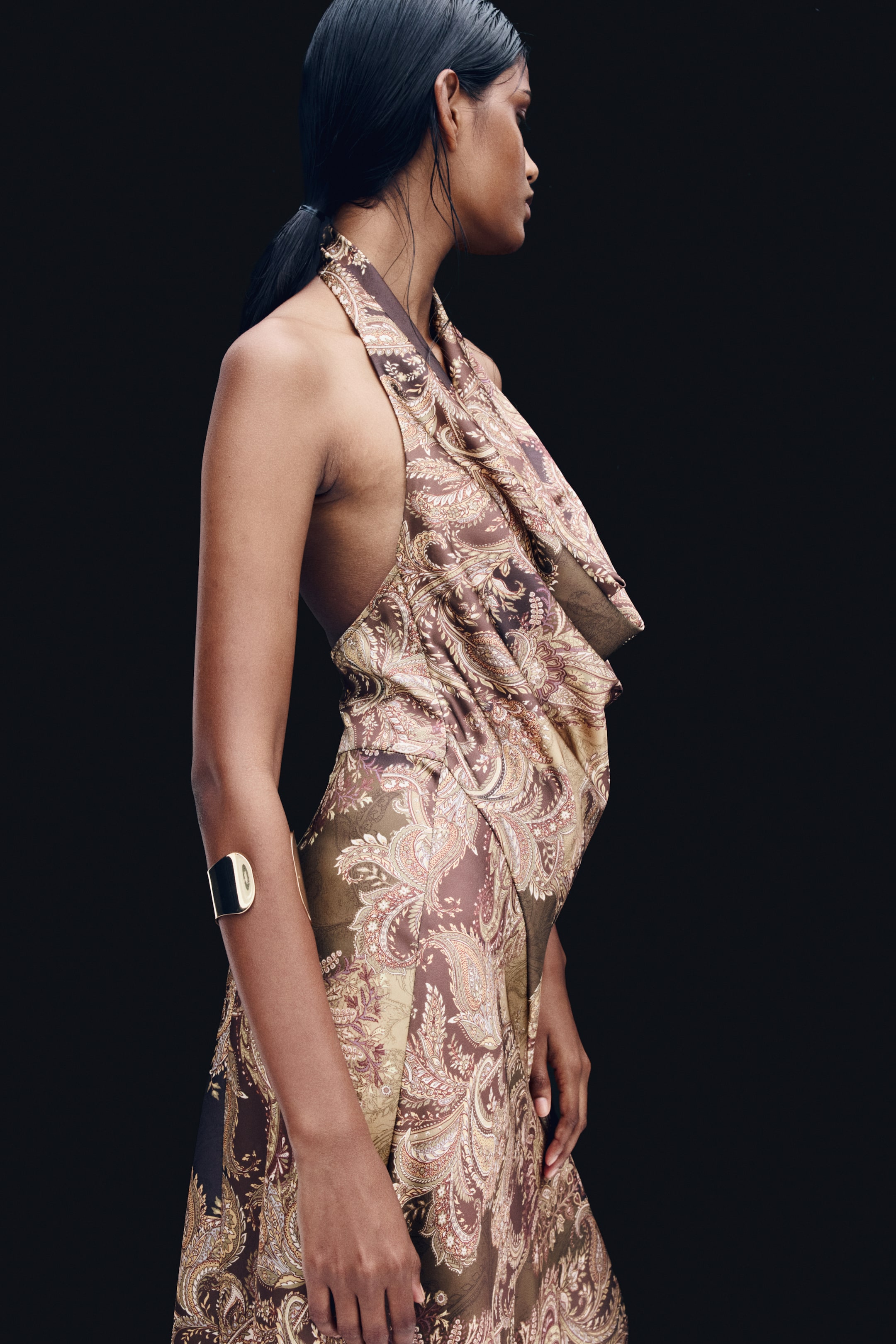 View larger image: A halterneck, paisley-patterned dress with a deep, draped cowl neckline and a subtle sheen is being worn by the model. The dress is shown from a side angle, highlighting its backless design and styled with a gold cuff bracelet on the model's arm.