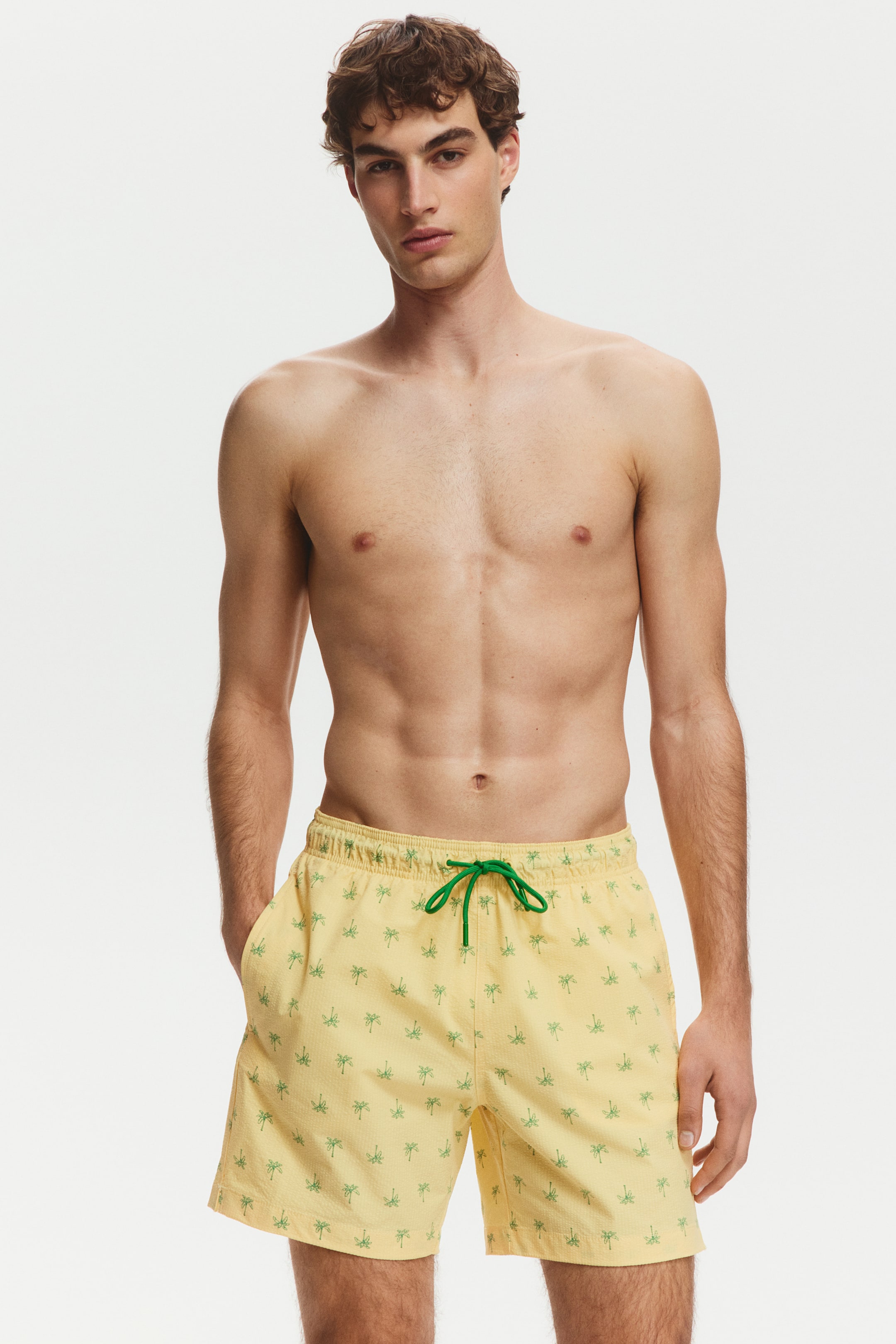 View larger image: Men's yellow swim trunks showcase a green palm tree print, an elastic waistband with a vibrant green drawstring, and side pockets, displayed from a front view.