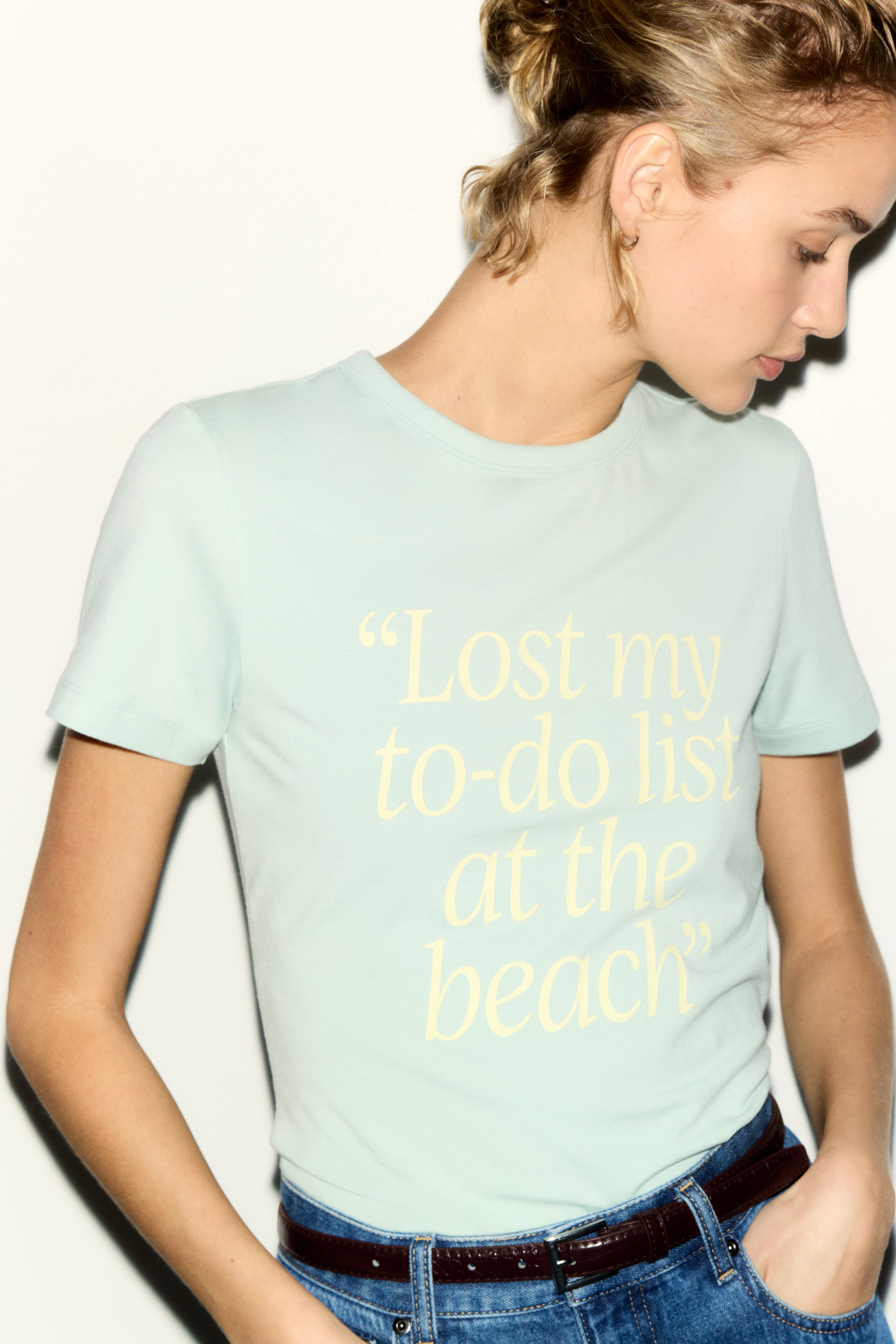 Se større billede: En lysegrøn kortærmet t-shirt med rund hals, der har den gule tekst "Lost my to-do list at the beach" på forsiden, bæres nede i blå jeans.