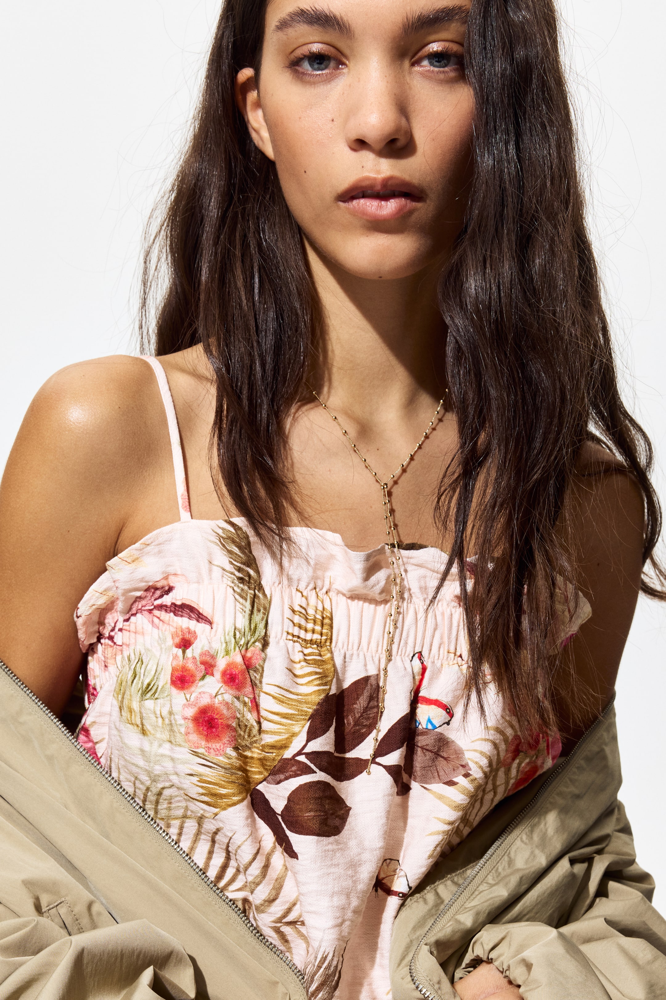 View larger image: The model is showcasing a strappy top adorned with frill trims and a vibrant pink tropical print. Complementing the top is a gold Y-necklace elegantly layered over it. Draped casually around the model's shoulders is a khaki jacket, adding a chic touch to the ensemble.