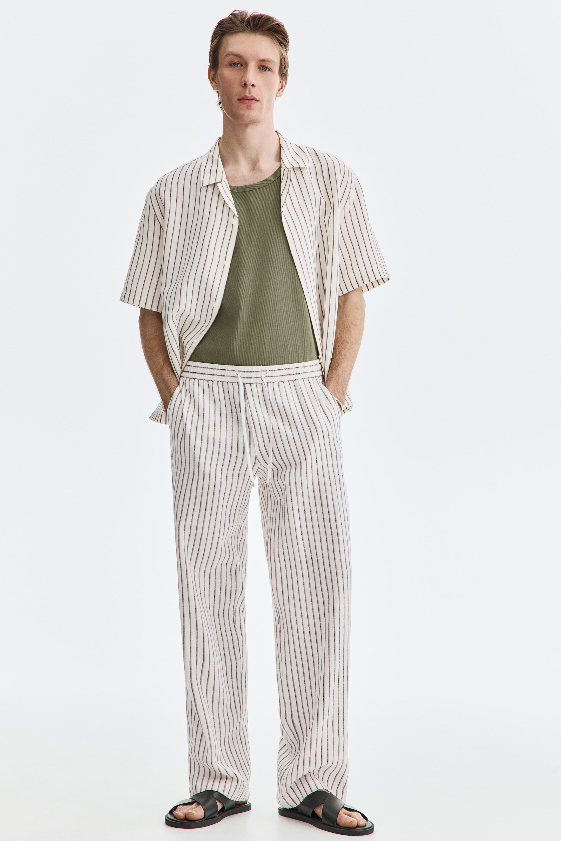 Ingrandisci l'immagine: Una camicia a maniche corte e pantaloni lunghi abbinati, entrambi caratterizzati da strisce verticali bianche e marrone scuro, sono presentati da un'angolazione frontale. La camicia, con colletto cubano, è indossata aperta sopra una t-shirt verde oliva scuro, mentre i pantaloni a gamba dritta presentano una vita con coulisse e sono abbinati a sandali neri.