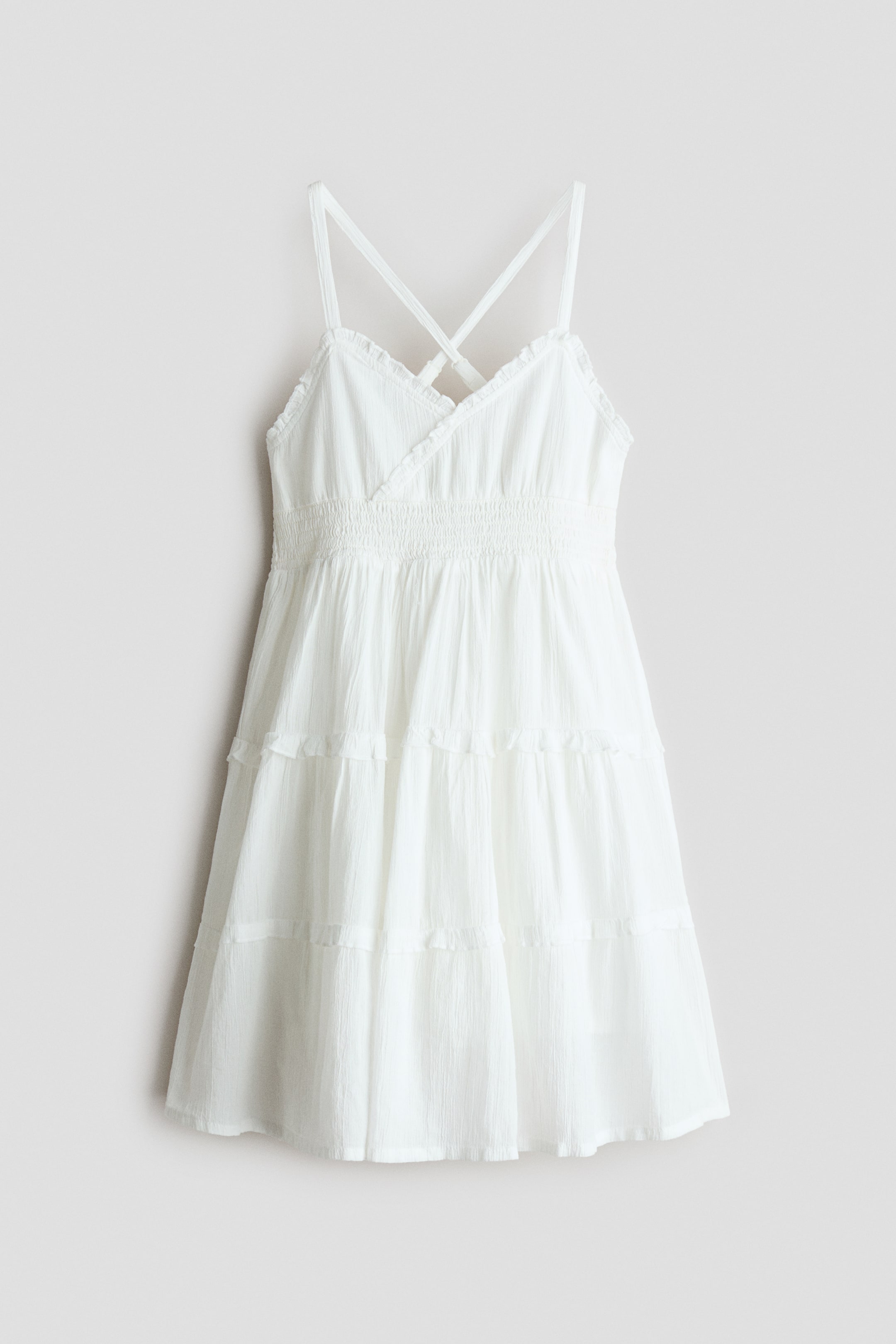 View larger image: A white tiered dress showcasing a crinkled texture, a V-neckline adorned with delicate ruffle trim, and slender spaghetti straps that cross at the back. It features a smocked elastic waist and a full tiered skirt with gathered seams, presented FRONT.