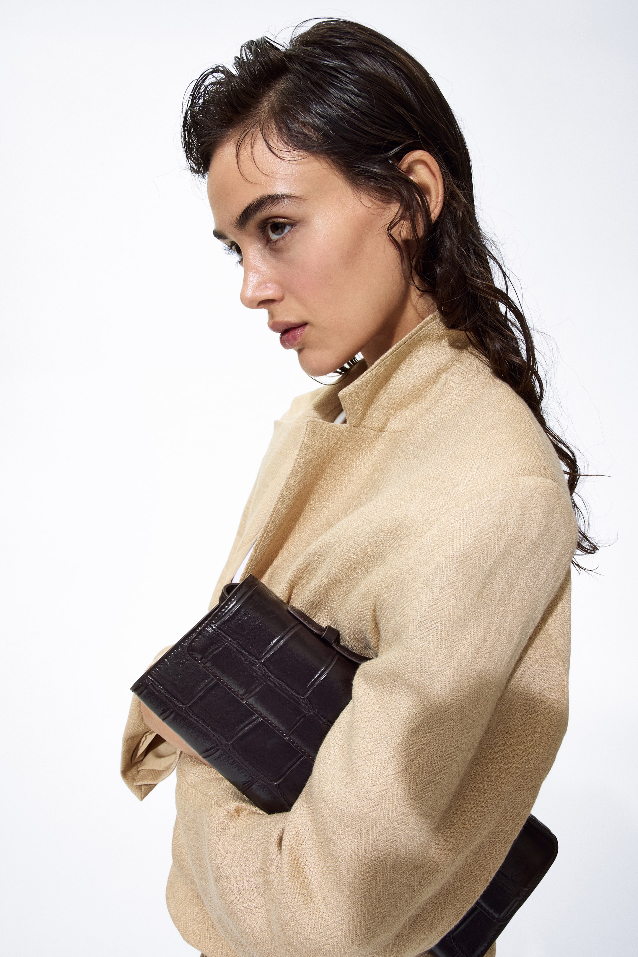 Ver imagen más grande: Un bolso de mano con grabado de cocodrilo en color marrón oscuro se sostiene ligeramente inclinado hacia adelante contra una chaqueta beige. Presenta un cierre de solapa prominente y una forma rectangular estructurada.