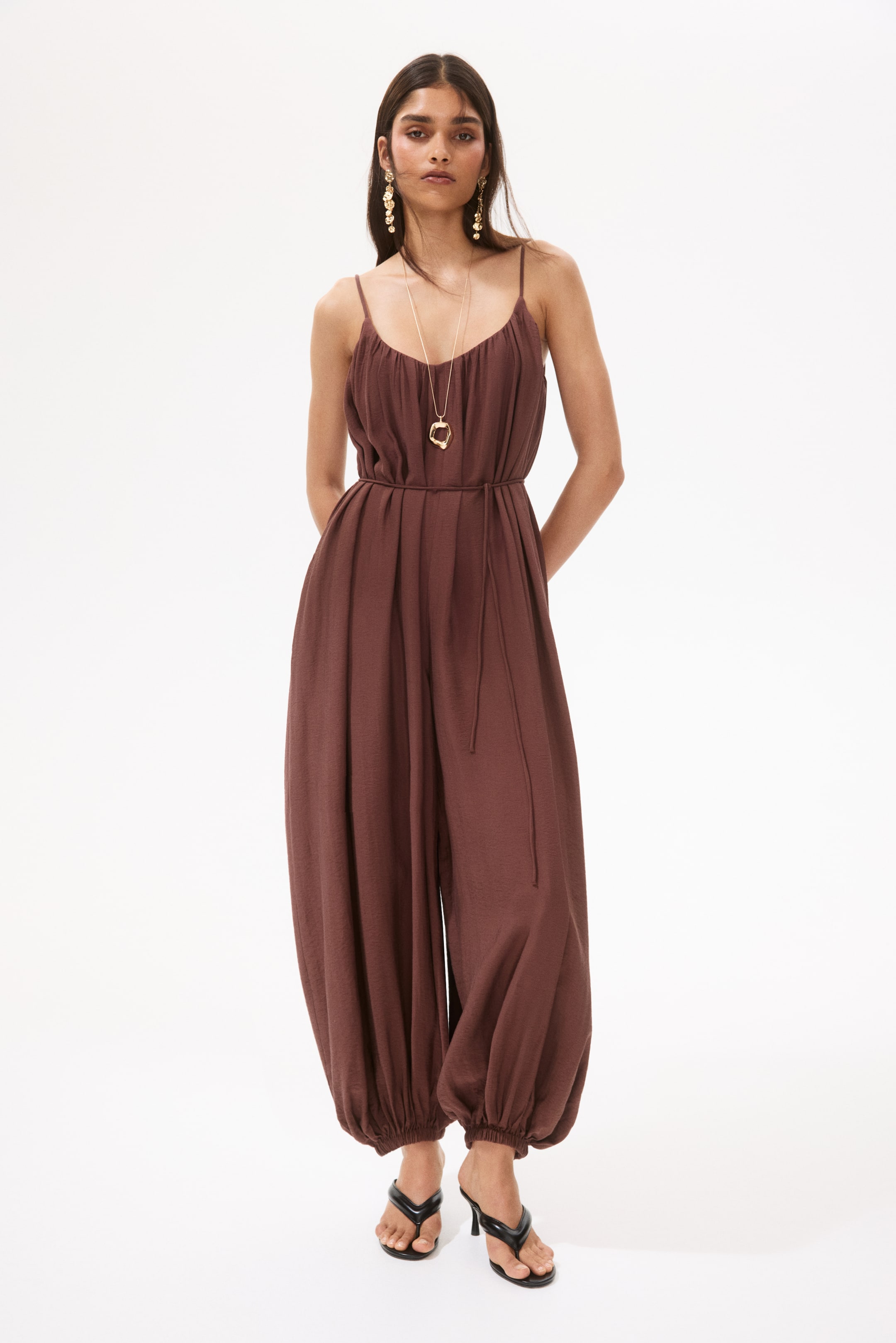View larger image: A dark brown jumpsuit is displayed front-facing, showcasing slender spaghetti straps and a ruched bodice. It's cinched at the waist with a tie, forming pleats, and flows into loose, balloon-style trousers gathered at the ankles. This ensemble is paired with black high-heeled thong sandals.