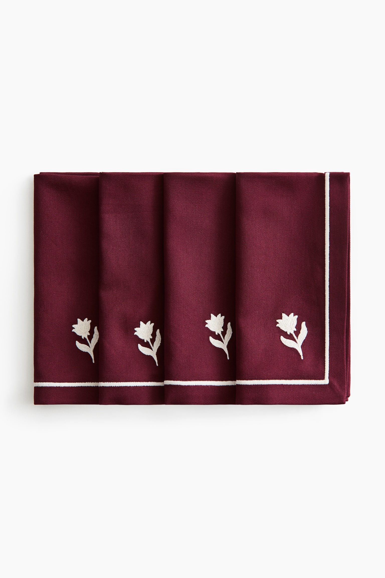 Four folded burgundy cloth napkins are arranged in an overlapping row, facing forward. Each napkin features a small white embroidered flower above a thin white embroidered border running along the bottom hem.