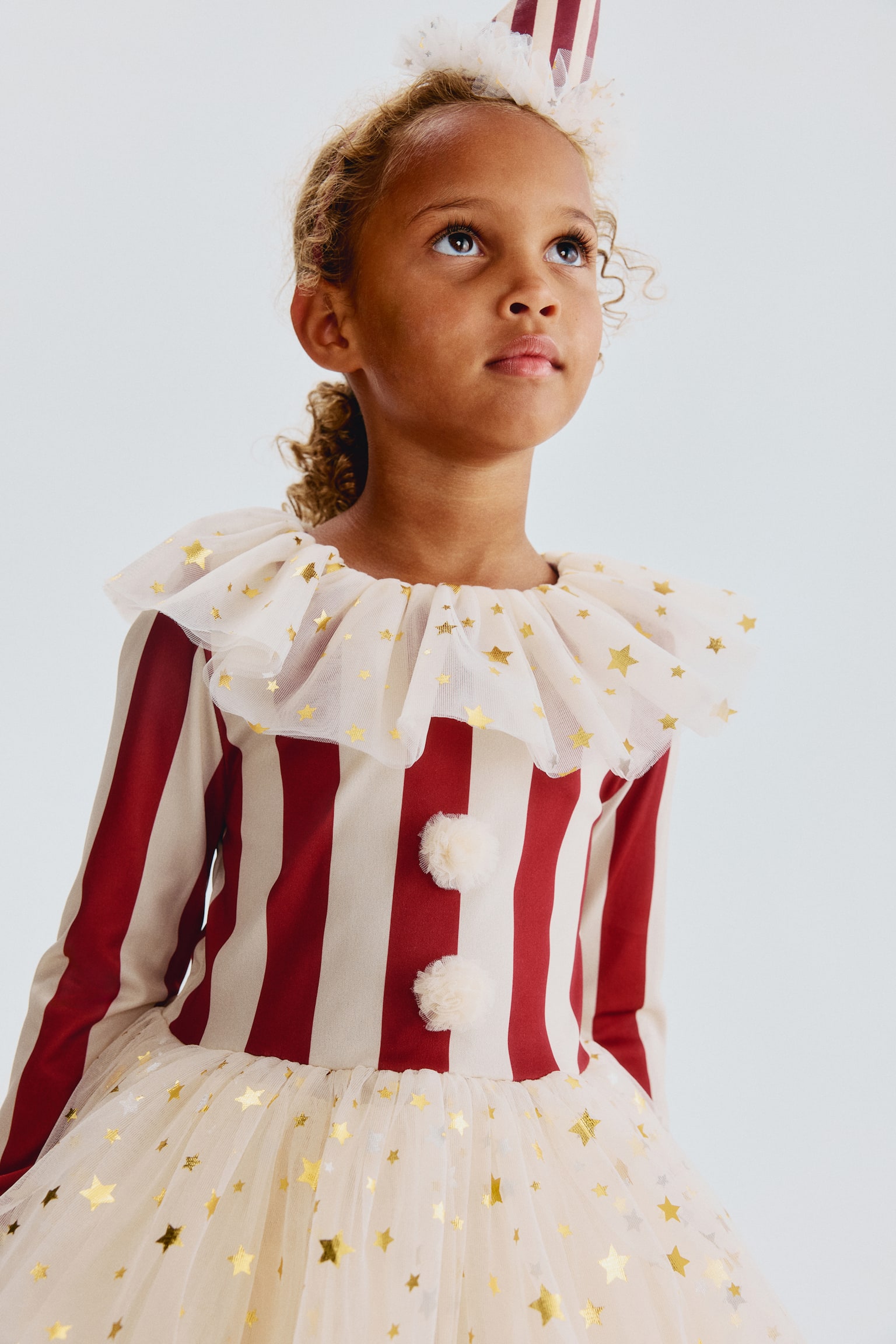 A childrenâs dress, shown from the front, featuring a burgundy and cream vertically striped bodice with long sleeves and ruffled tulle cuffs. The dress showcases a layered, ruffled tulle collar and a matching tulle skirt, both adorned with gold foil star prints, along with two fluffy cream pompoms on the chest. - 1