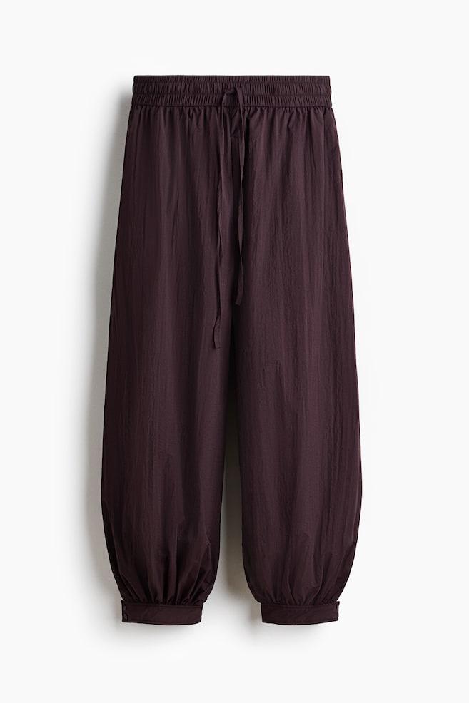 Deep plum, ankle-length trousers showcase their front view, featuring an elasticated, drawstring waist and discreet side pockets. They flaunt voluminous balloon legs, gathering into cuffed hems with visible buttons. - 1