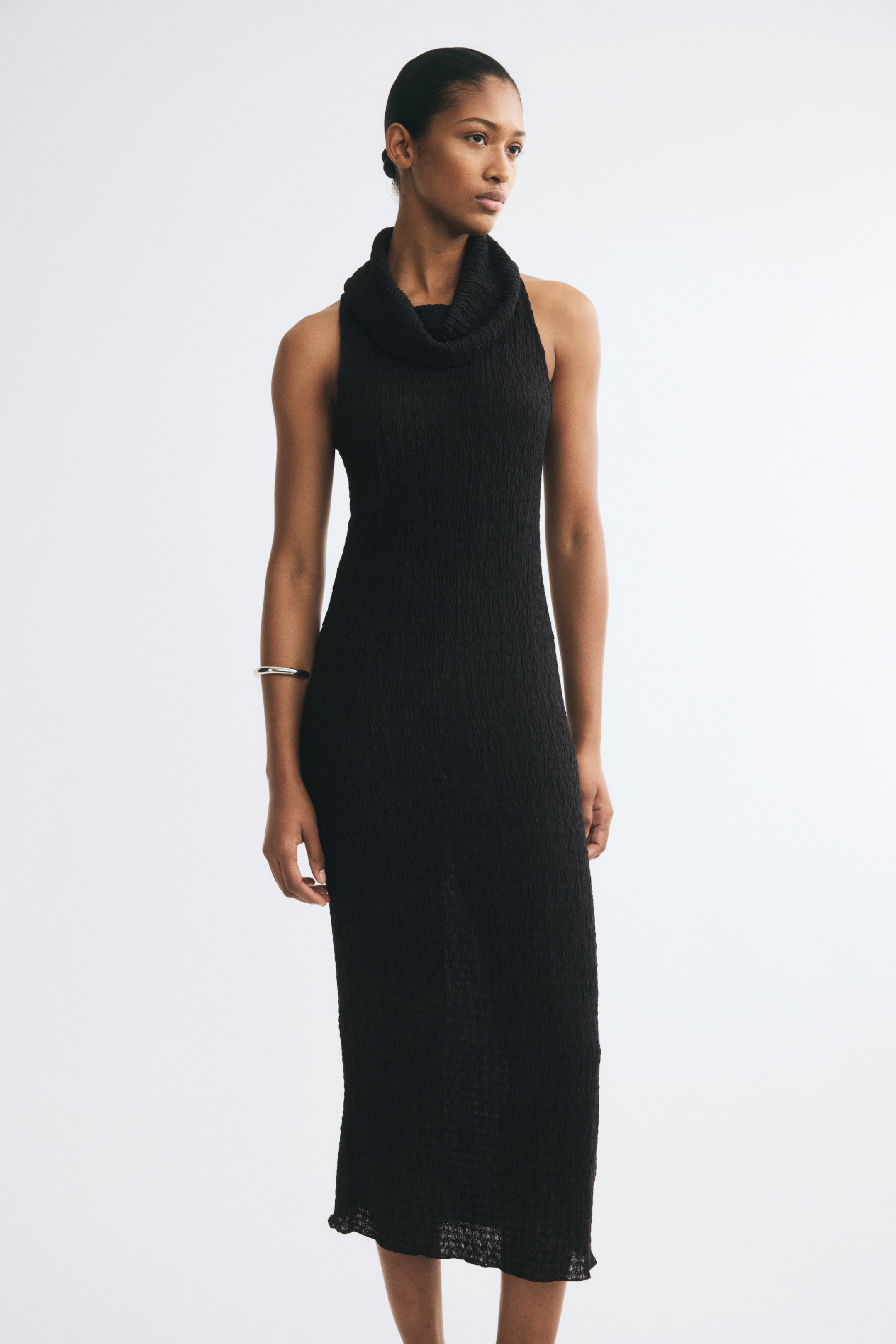View larger image: The model is wearing a black, calf-length sleeveless dress made from crinkled jersey fabric. The dress features a slim fit, a high foldover cowl neckline, and a textured finish.