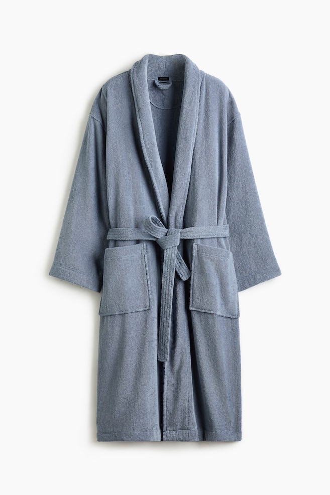 A blue-grey dressing gown in soft cotton terry is presented facing front, featuring a shawl collar, two front pockets, and a detachable tie belt tied at the waist. - 1