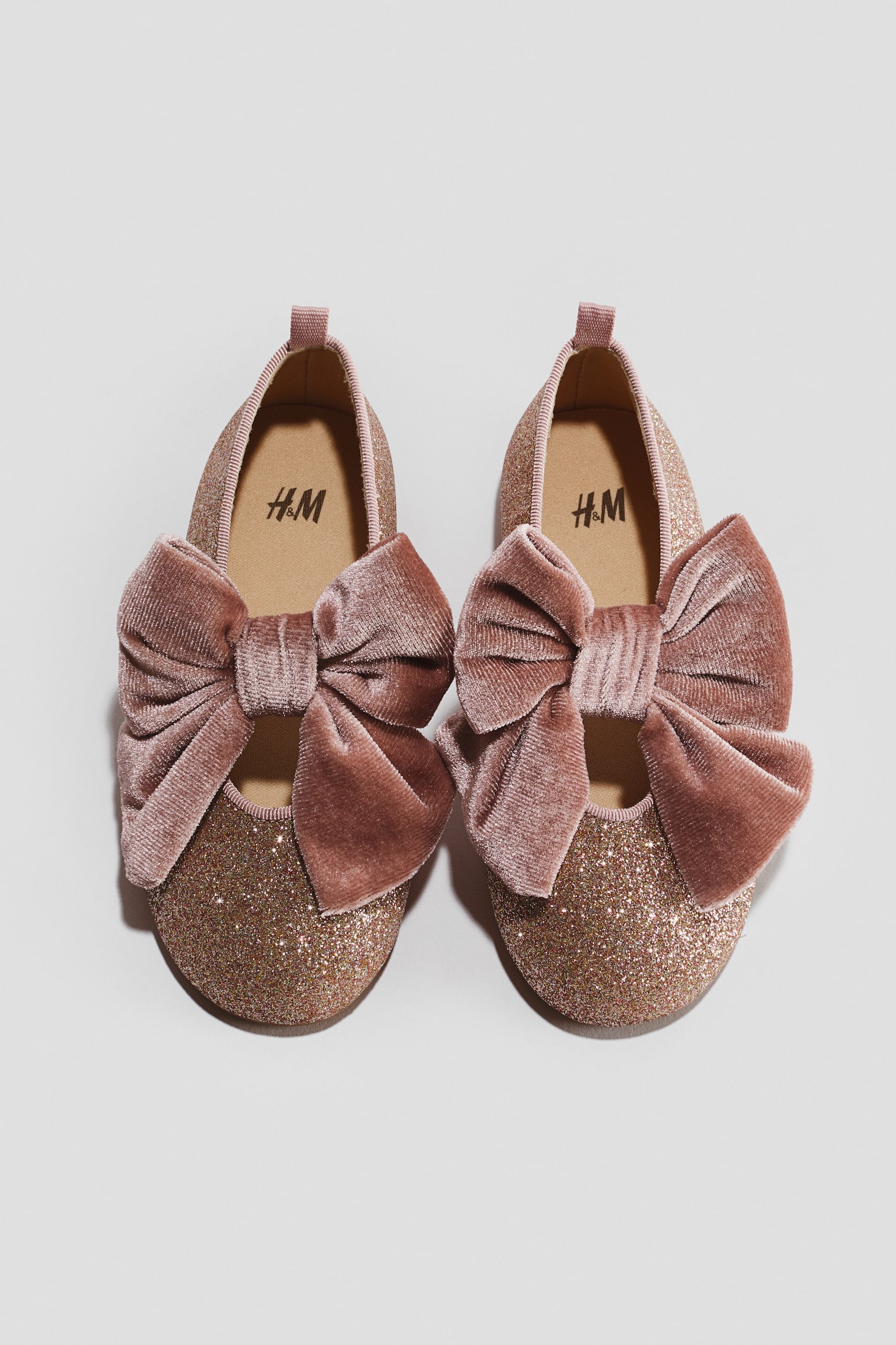 A pair of children's slip-on shoes is facing front, showcasing a glittery rose-gold finish on the uppers. Each shoe is featuring a large, dusty pink velvet bow across the instep and is displaying a small heel pull tab at the back.