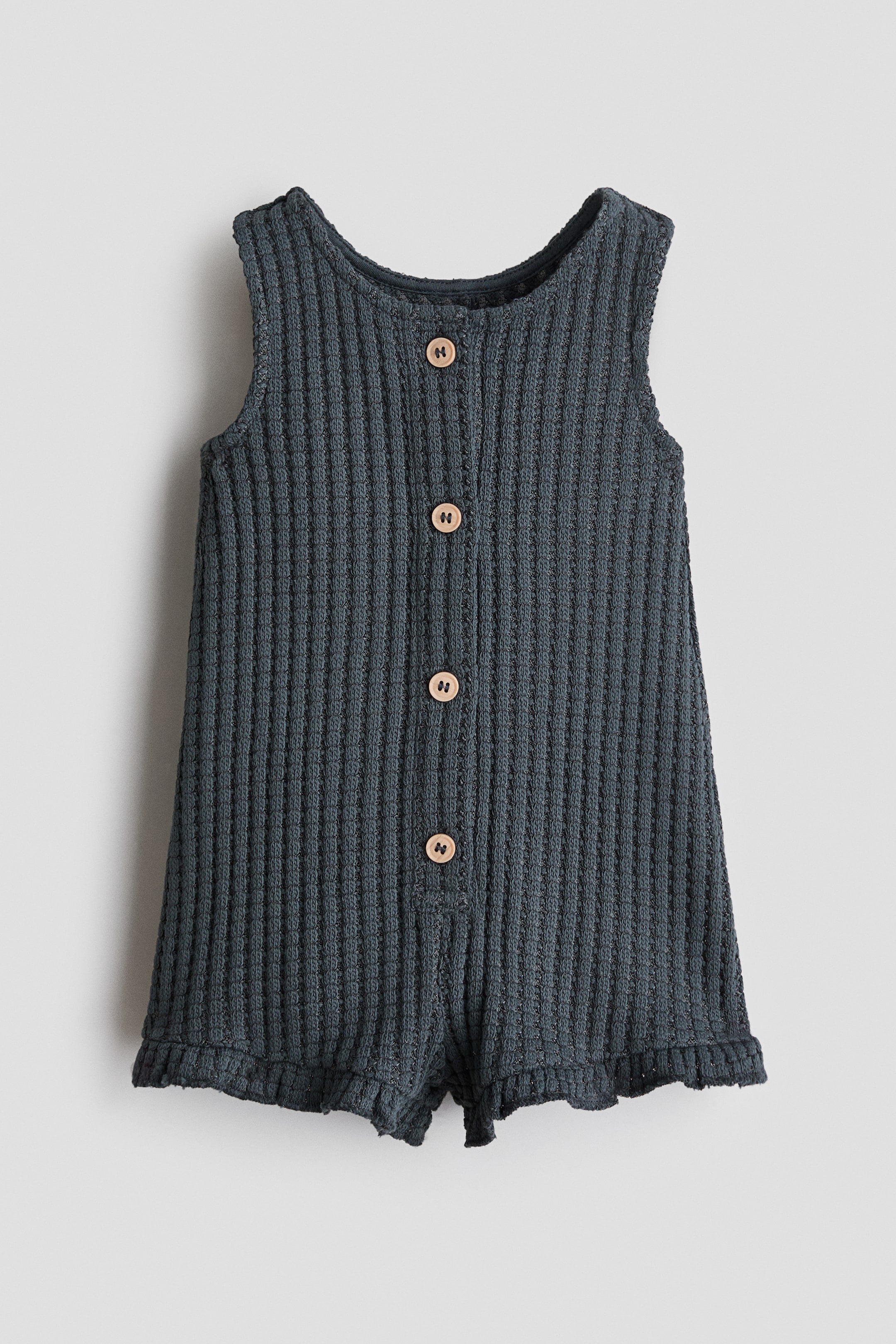 View larger image: "A dark grey sleeveless romper suit is showcased from the front, crafted from a soft waffle-knit fabric. It features a round neckline, wooden buttons running down the front, short legs, and a narrow frill trim at the hems."