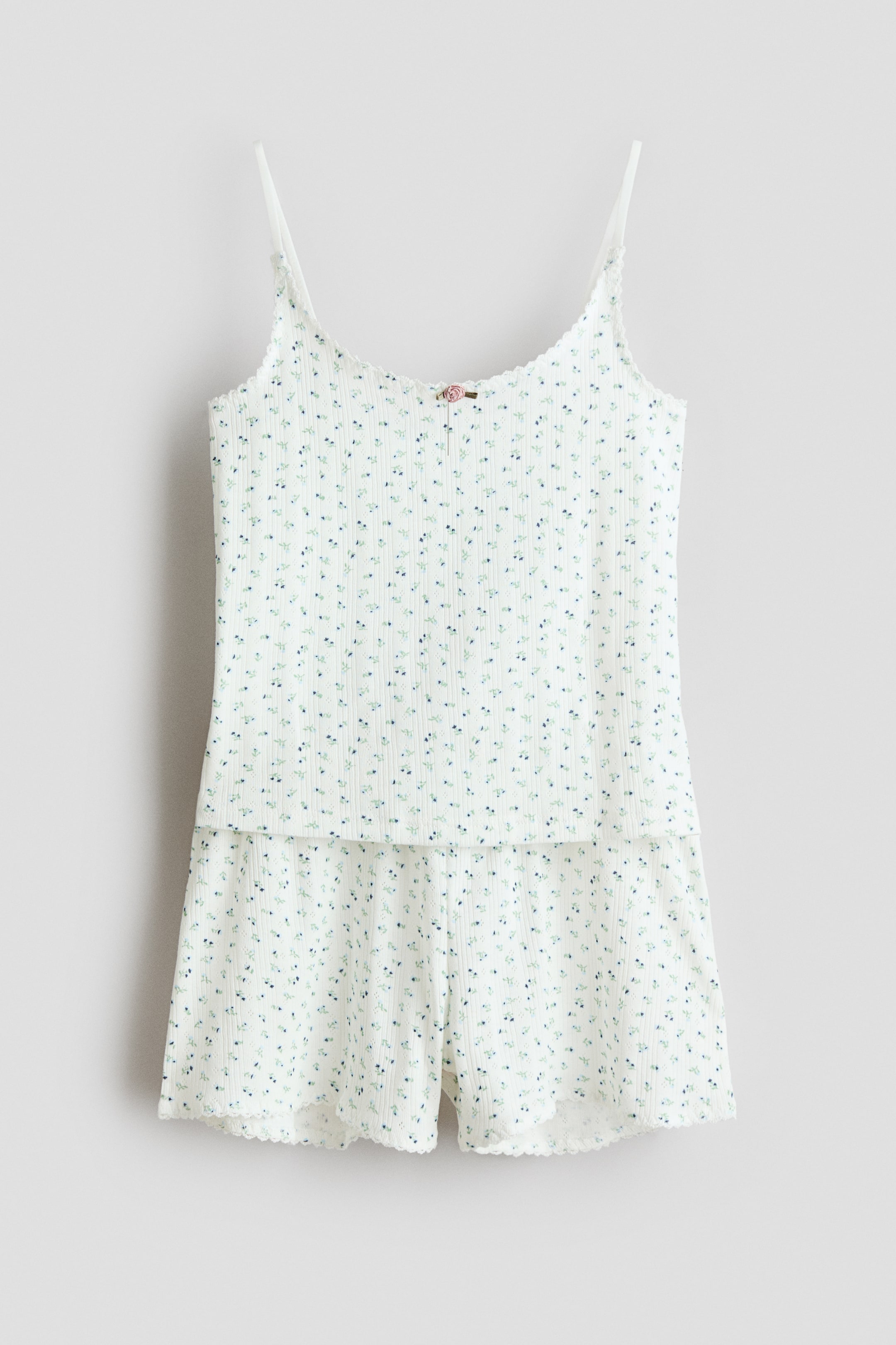 View larger image: A two-piece pyjama set is visible from the front, consisting of a white ribbed camisole top and matching shorts. The set features a small blue and green floral print, with scalloped trim outlining the top's neckline and the shorts' leg openings, while a small pink rose decorates the centre front of the top.
