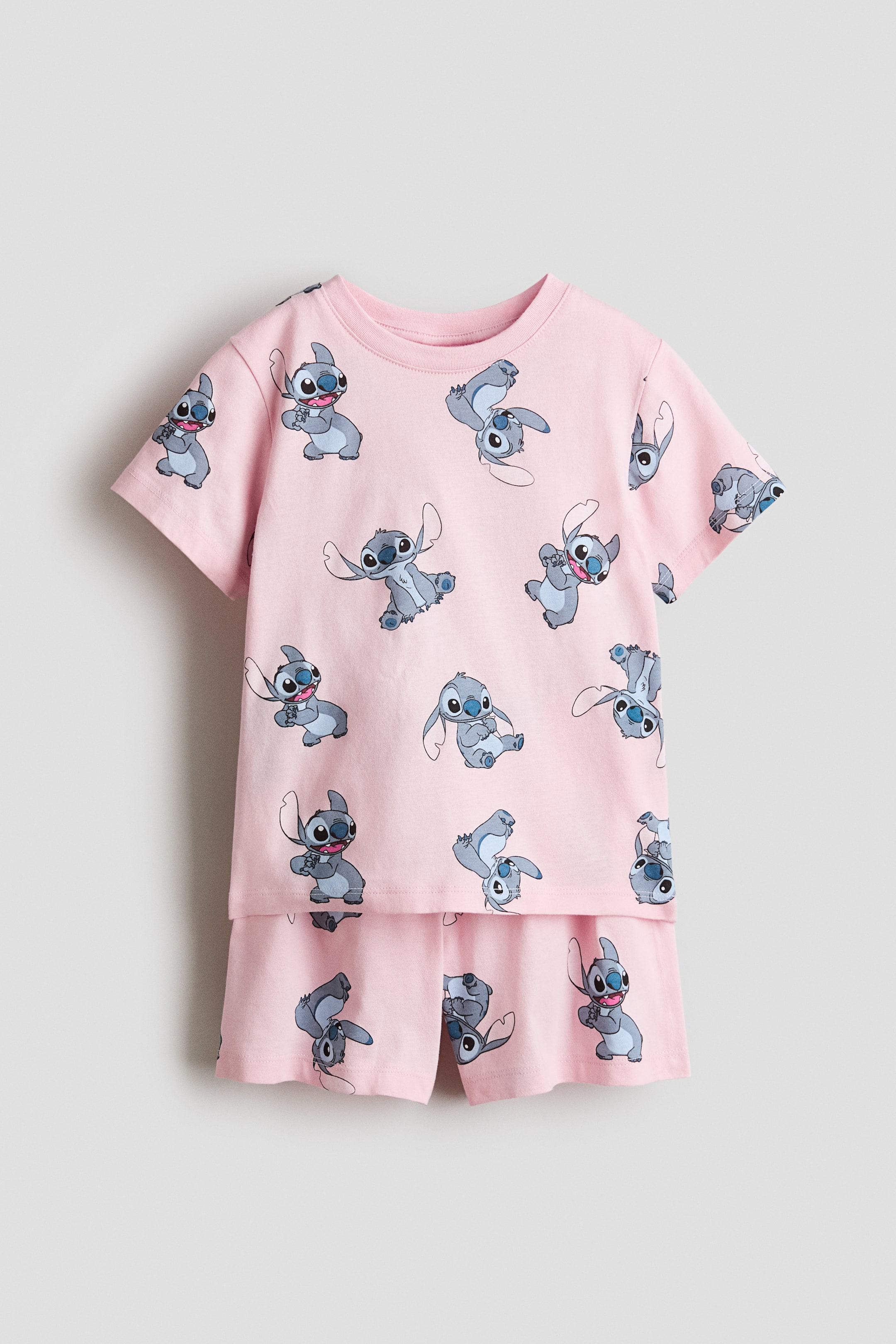 Ver imagem maior: Um conjunto de pijama rosa é apresentado de frente, composto por uma peça superior de mangas curtas com gola redonda e calções a condizer, todos exibindo uma estampa integral do Stitch em várias poses.