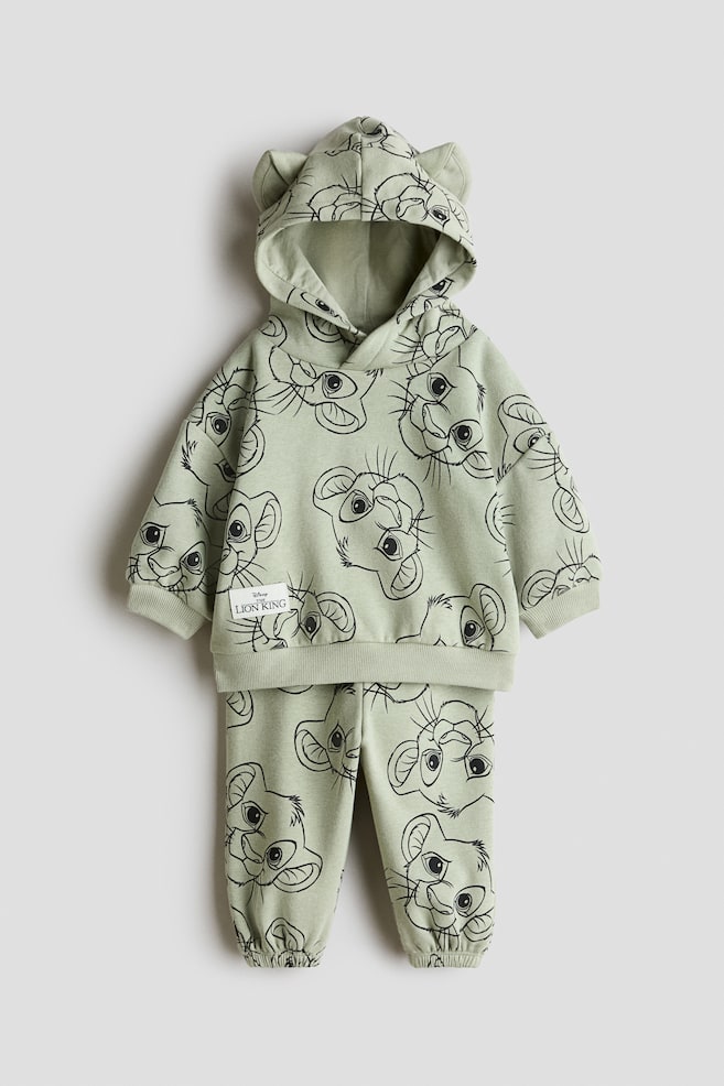 Cet ensemble sweat à capuche et pantalon de jogging, présenté de face, se compose dâun tissu vert clair orné dâun imprimé intégral noir en contour de Simba, tiré de The Lion King de Disney. Le sweat à capuche présente une capuche avec des oreilles en tissu attachées, des épaules tombantes ainsi que des poignets et un ourlet côtelés, tandis que le pantalon de jogging assorti est doté de poignets froncés élastiqués. - 1