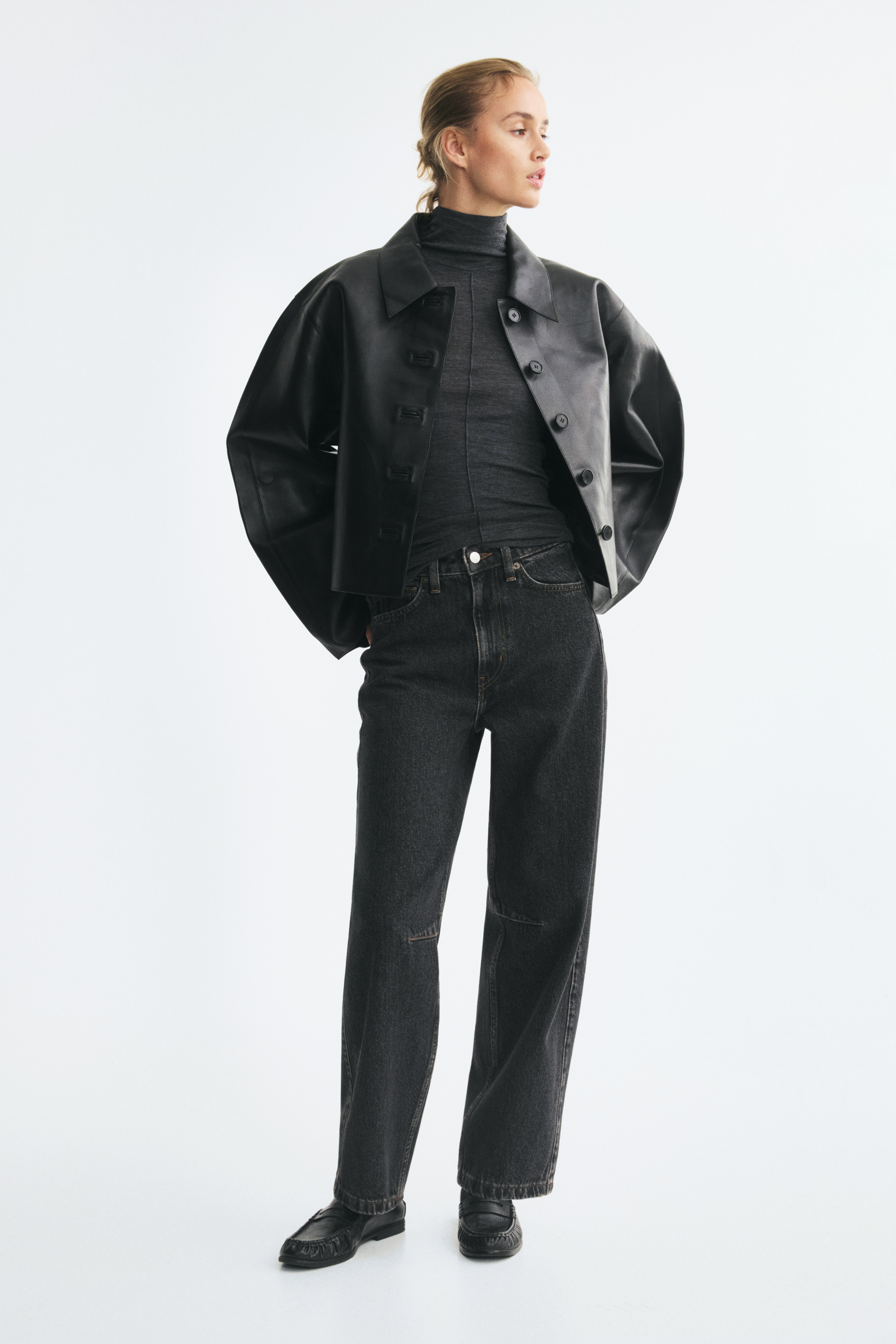 A pair of high-waisted, five-pocket jeans in dark grey washed denim is hanging flat in a front view, showing a curved, voluminous leg silhouette that tapers towards the hem. The design features a classic zip fly with a silver-tone button fastening and horizontal seam detailing at the knees.
