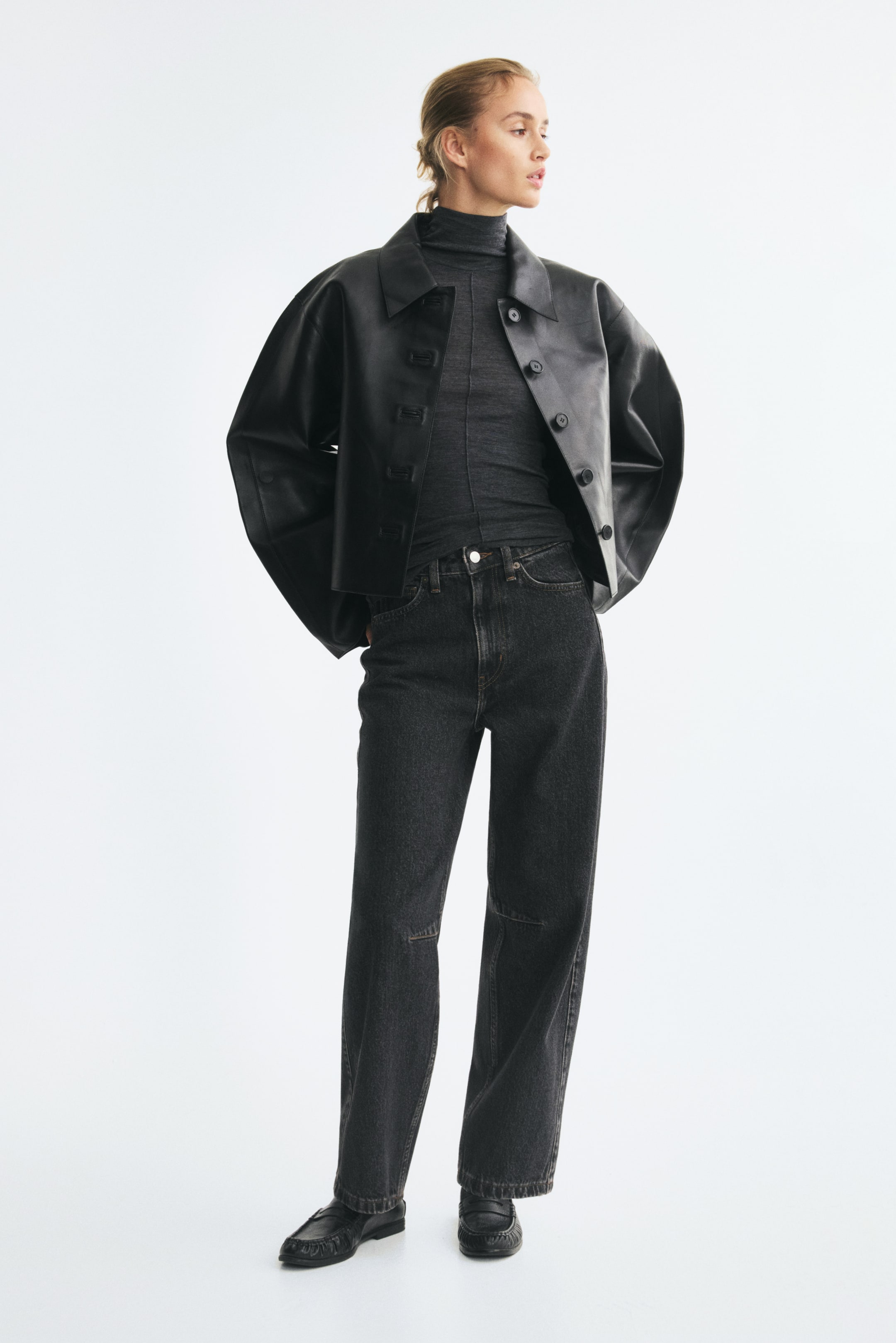 Se større billede: Mørkegrå vaskede denimjeans med høj talje, løs pasform og voluminøse, buede ben, der smalner ved kanten, bæres af modellen. Jeansene styles med en nedstoppet mørkegrå rullekravebluse, en sort læderjakke og sorte loafers.