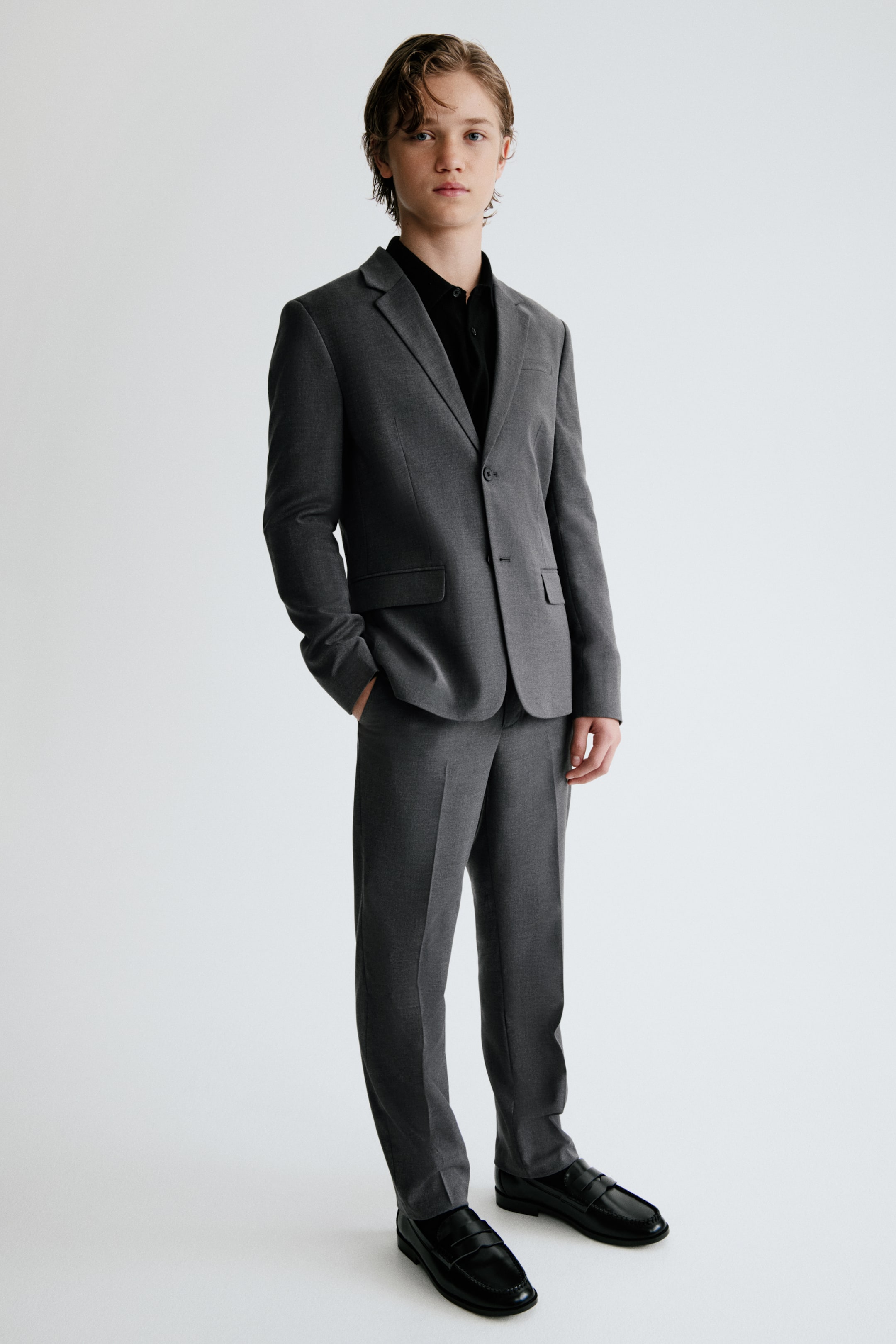Voir une image plus grande: Un complet en sergé extensible gris est présenté de face, mettant en valeur une veste structurée à revers crantés, une fermeture avant à deux boutons et des poches à rabat. Le pantalon tailleur assorti affiche des plis marqués à l’avant et des poches latérales en biais, agencé avec une chemise noire à col et des mocassins noirs.