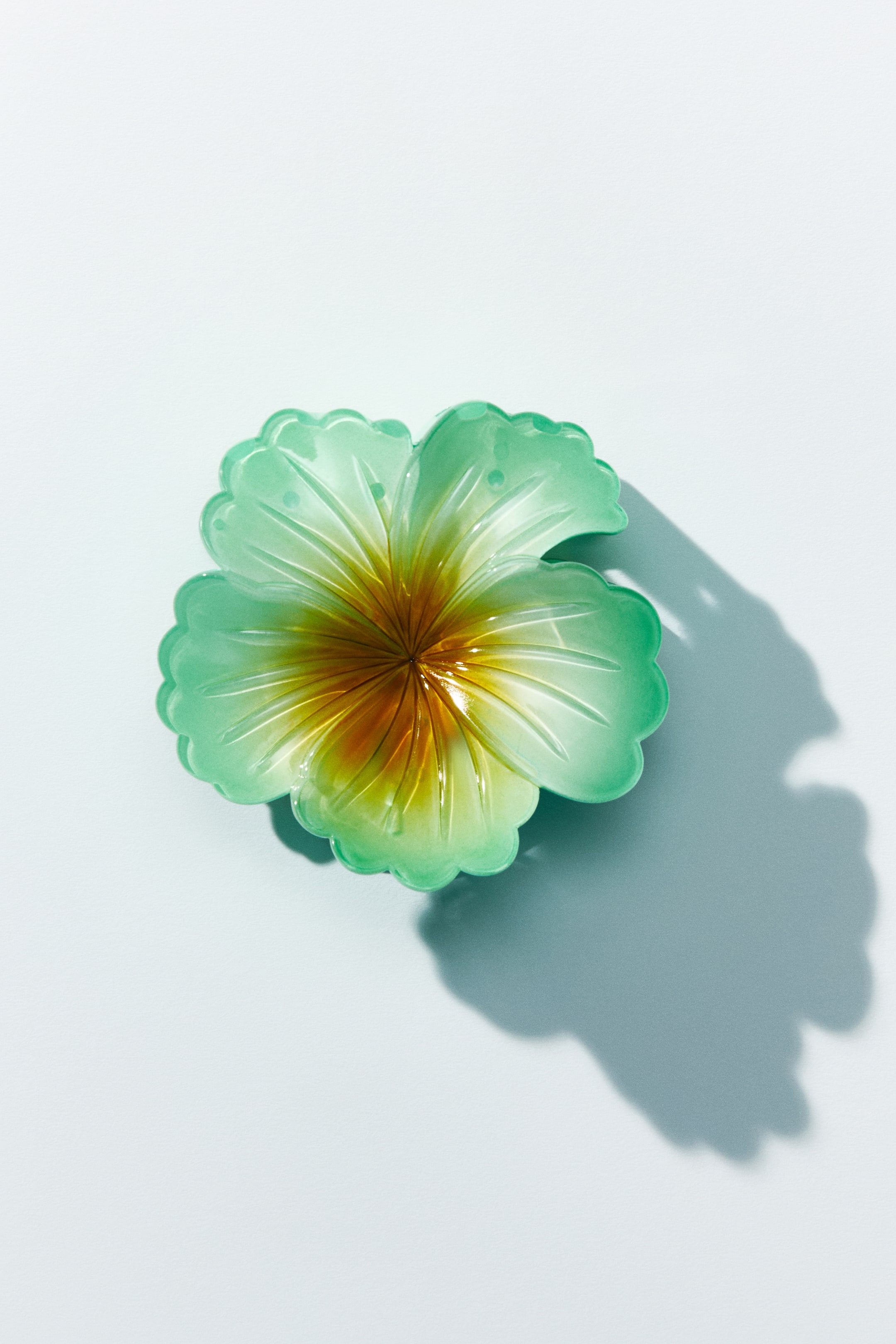 View larger image: A translucent flower-shaped hair claw featuring green scalloped petals transitioning to a yellow-orange centre. The petals display visible veins, and the claw is presented from a top-down angle.